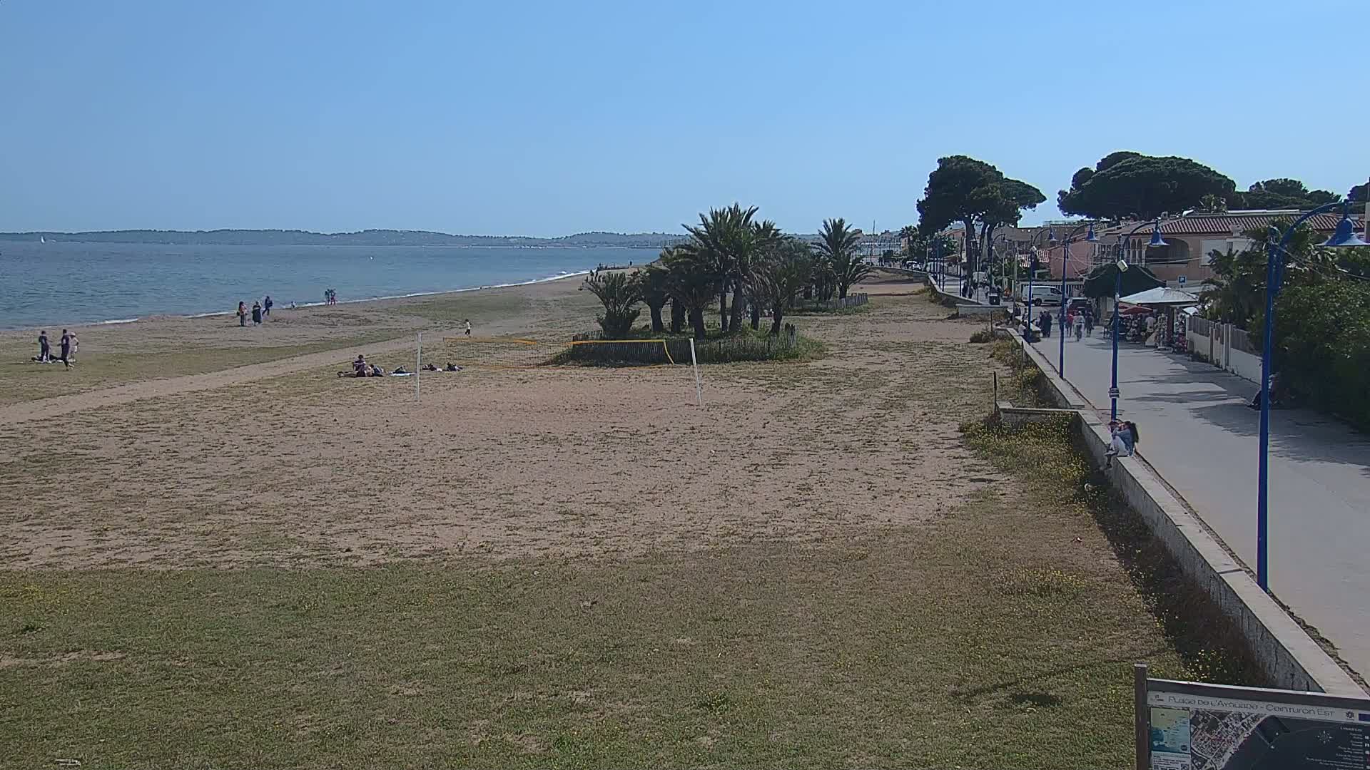 A sandy beach with sparse crowds, palm trees, and a paved walkway next to buildings stretches along a calm, blue body of water under a clear sky.