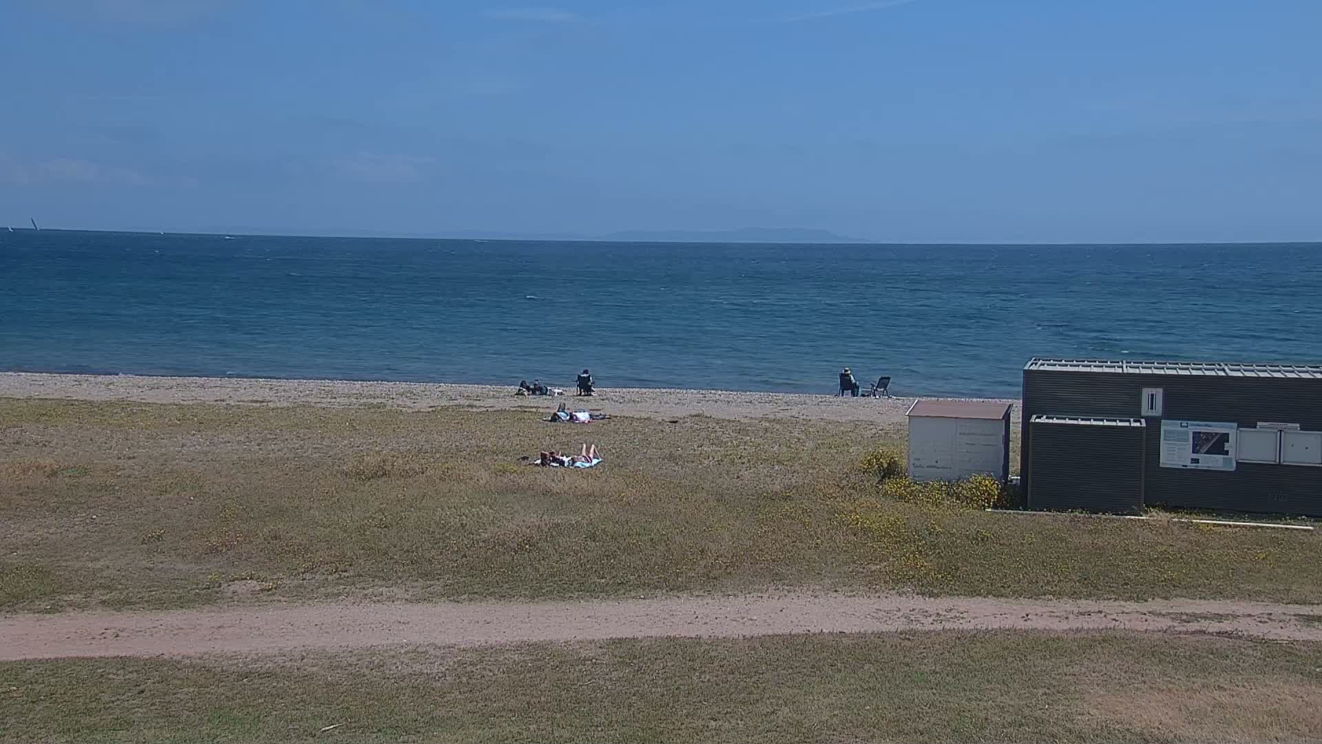 A mostly sunny day at a beach shows several people sunbathing and sitting on the sand near a small building, with a calm ocean in the background.