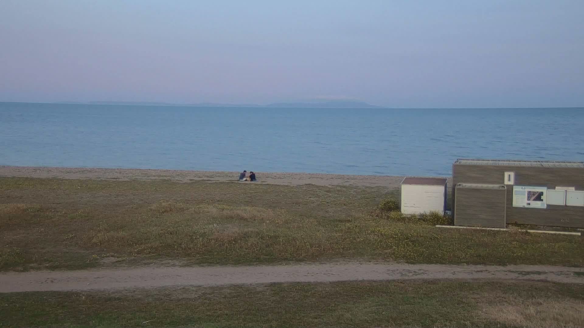 A calm, pale blue sea stretches to a distant horizon, where a faint line of land is visible; a sandy beach lies below, with two people sitting close together, and two small buildings stand to the right of the beach.