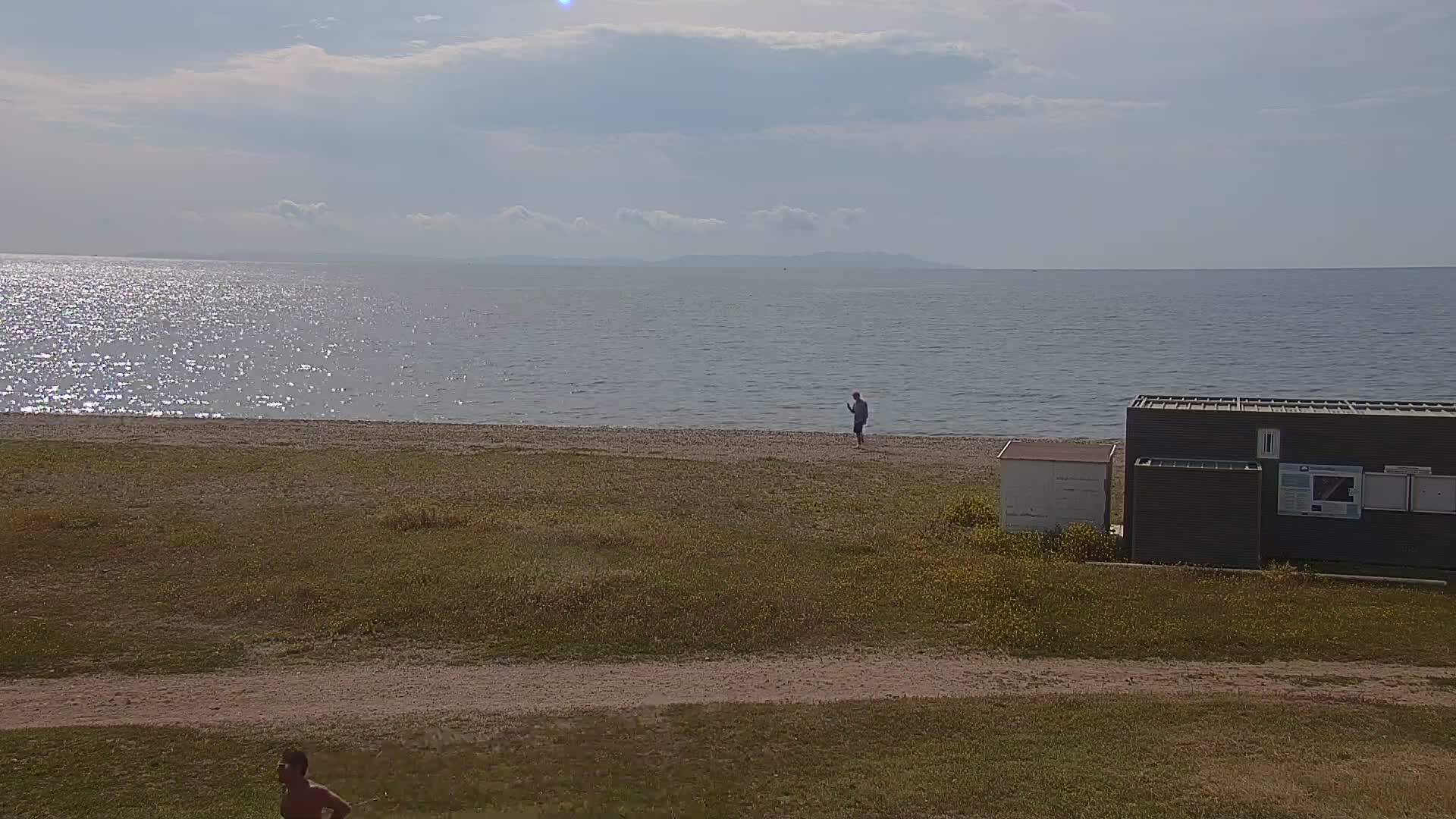 A mostly sunny day shows a calm sea with a person standing on a sandy beach near a small building and grassy area.