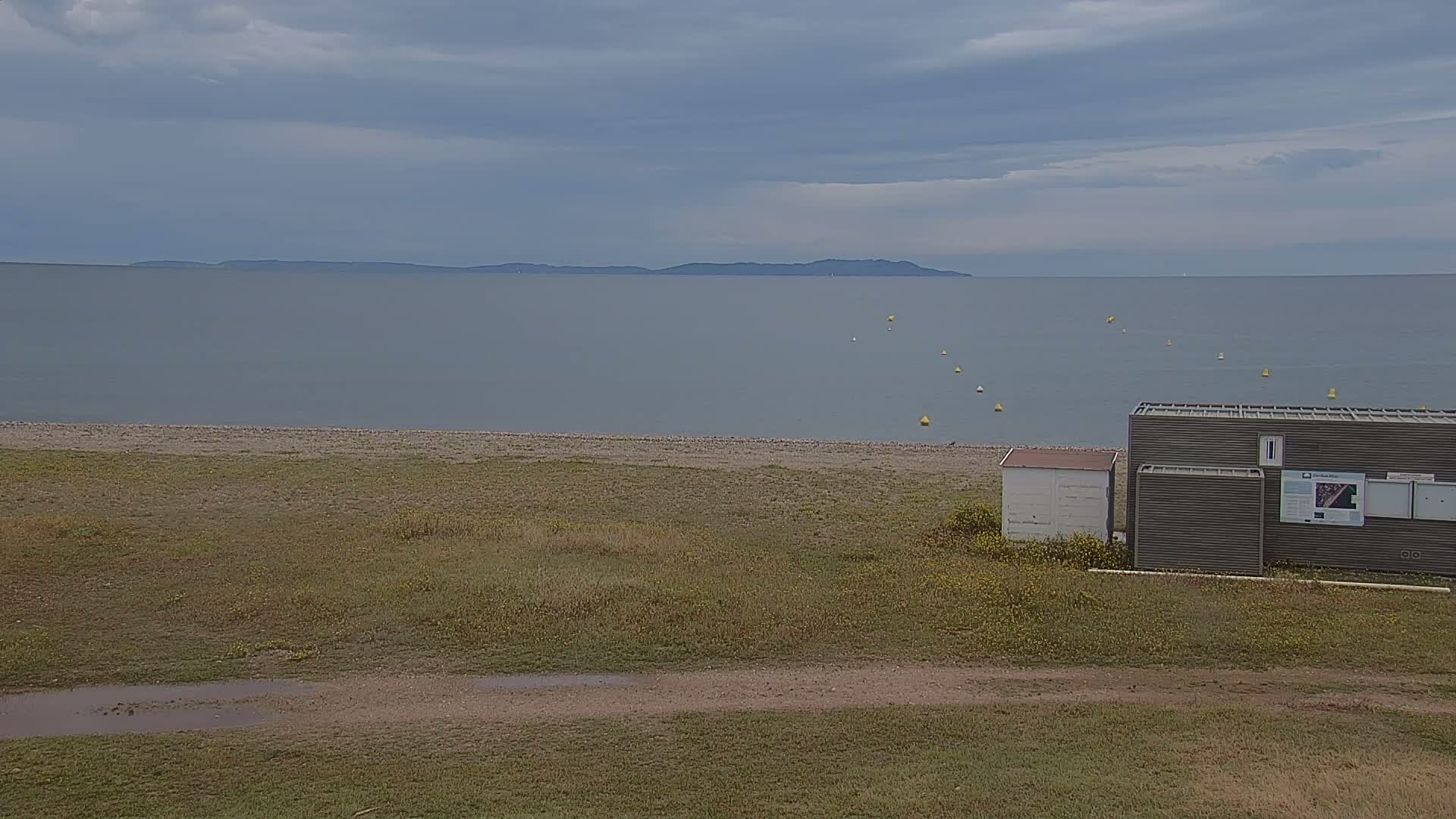 A calm, gray-skied day overlooks a beach with a small building and a distant island visible across a body of water.