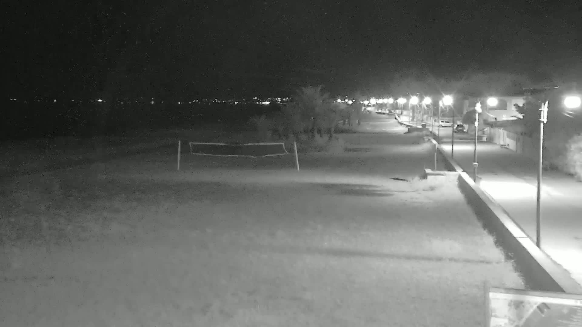 A nighttime, low-light view shows a beach with a volleyball net, a paved walkway lined with lights, and a few parked cars.