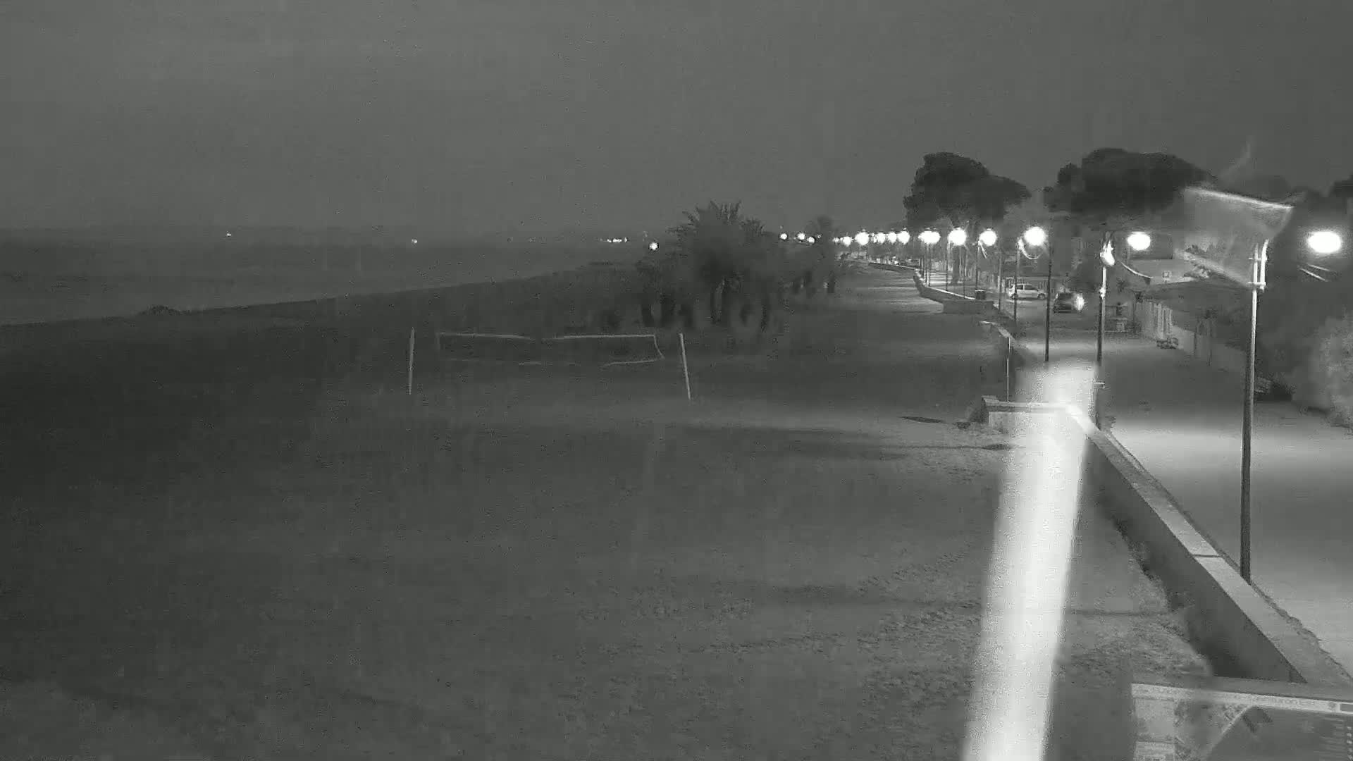 A dark, rain-swept beach at night is lit by a line of streetlights along a paved walkway bordering a row of palm and pine trees.