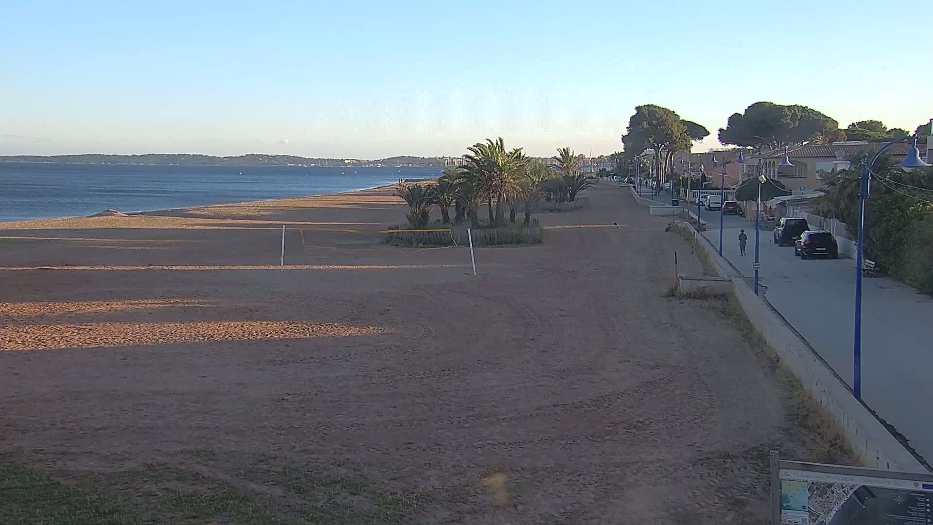 A sandy beach with a volleyball net, palm trees, and a paved walkway lined with buildings and cars extends to a calm, blue sea under a clear sky.