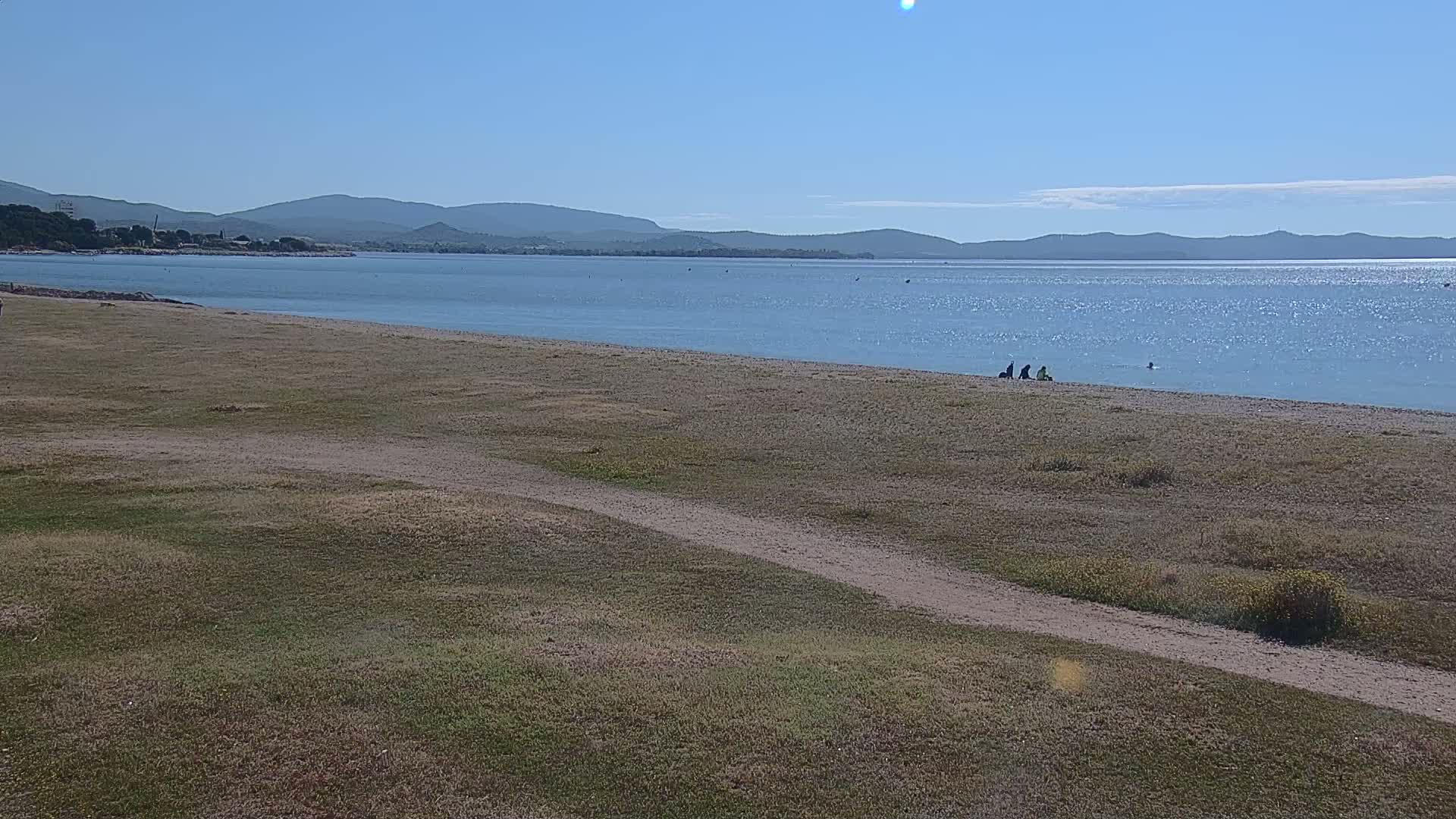 A calm, sunny day at a beach shows a few people sitting on the sand near a body of water with mountains in the distance.