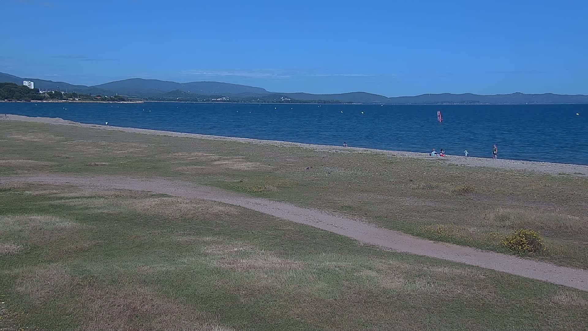 A sandy beach with a few people on it stretches along a calm blue sea under a clear blue sky, backed by a grassy area and distant mountains.