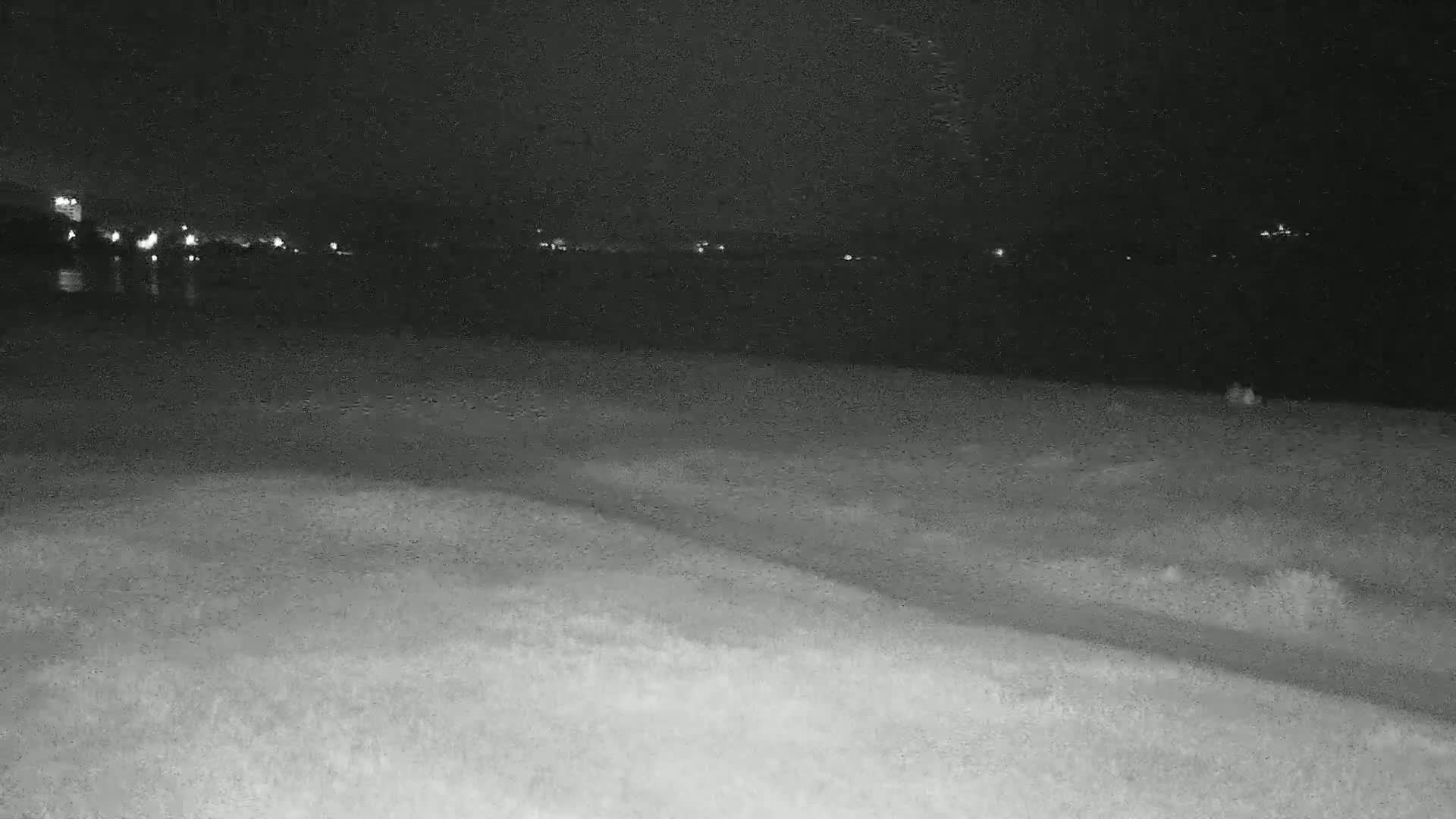 A nighttime, low-light view shows a dark body of water with distant lights, and a flat expanse of land in the foreground.