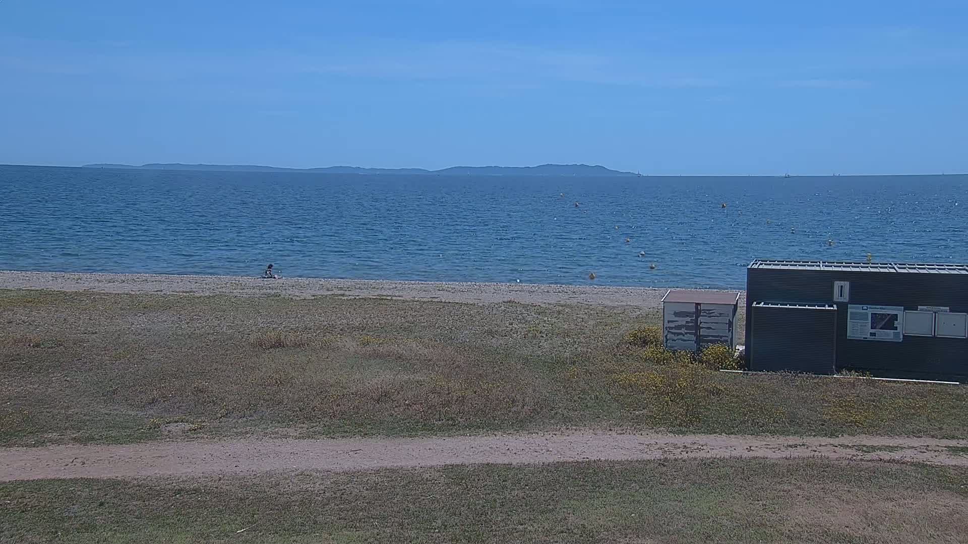 A sandy beach with a calm blue sea under a clear blue sky, a lone person sitting on the sand, and several small structures near the shore.