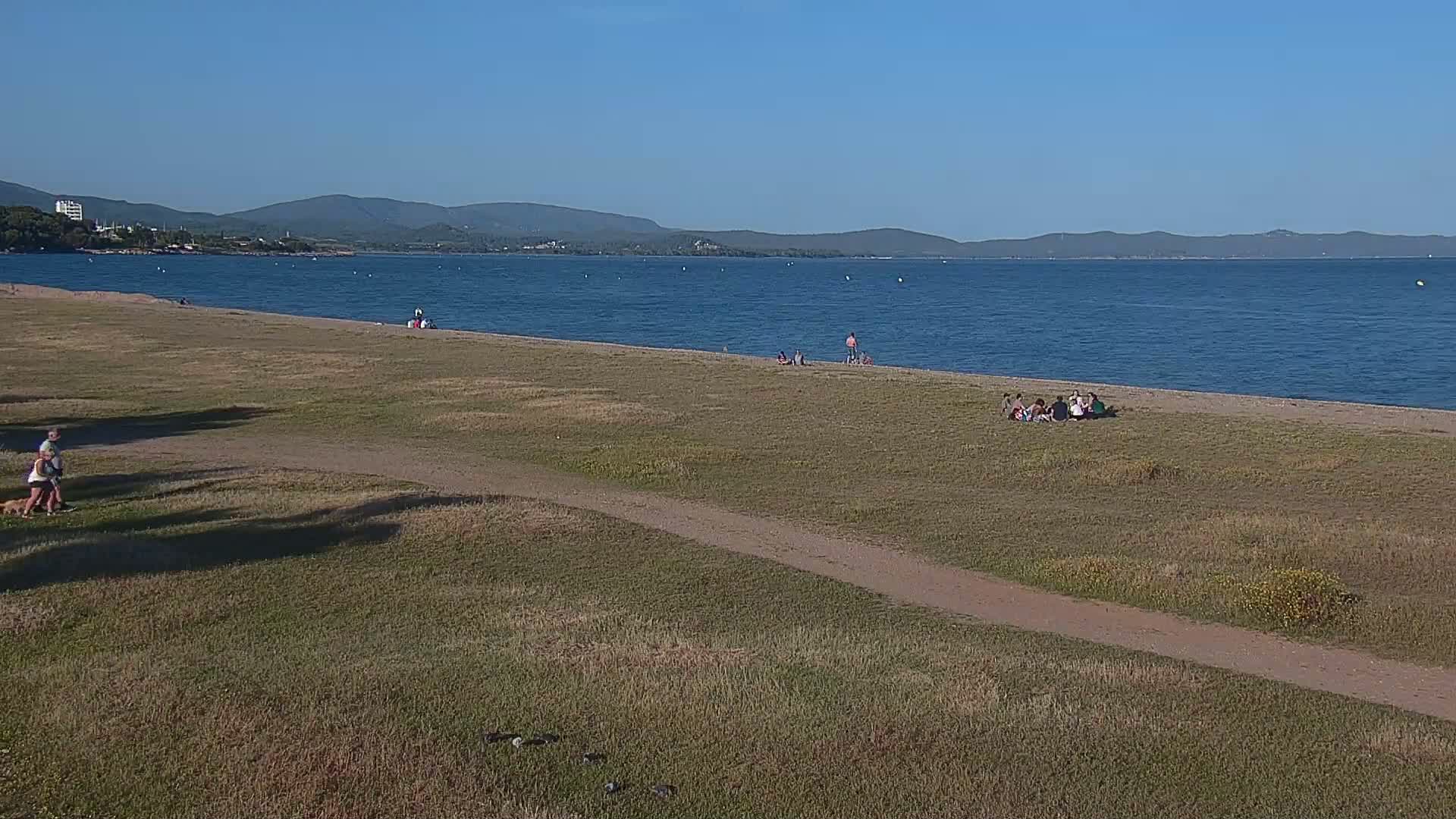 A sandy beach with scattered people sits beside a calm, blue body of water, backed by a grassy area and distant mountains under a clear blue sky.