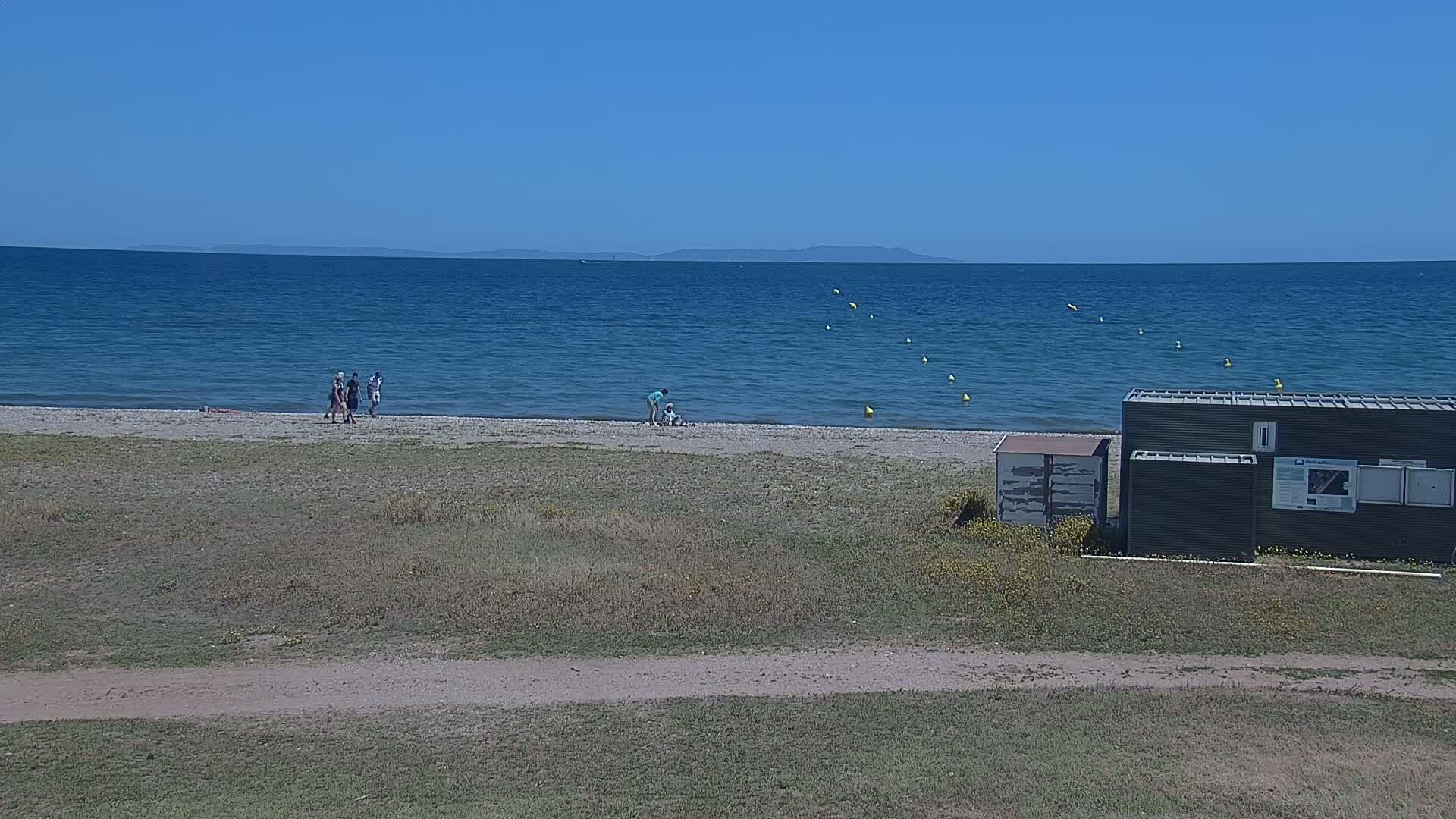 A sandy beach with a few people on it and a dark-colored building near the water's edge on a clear, sunny day, with a calm blue sea extending to a distant coastline.