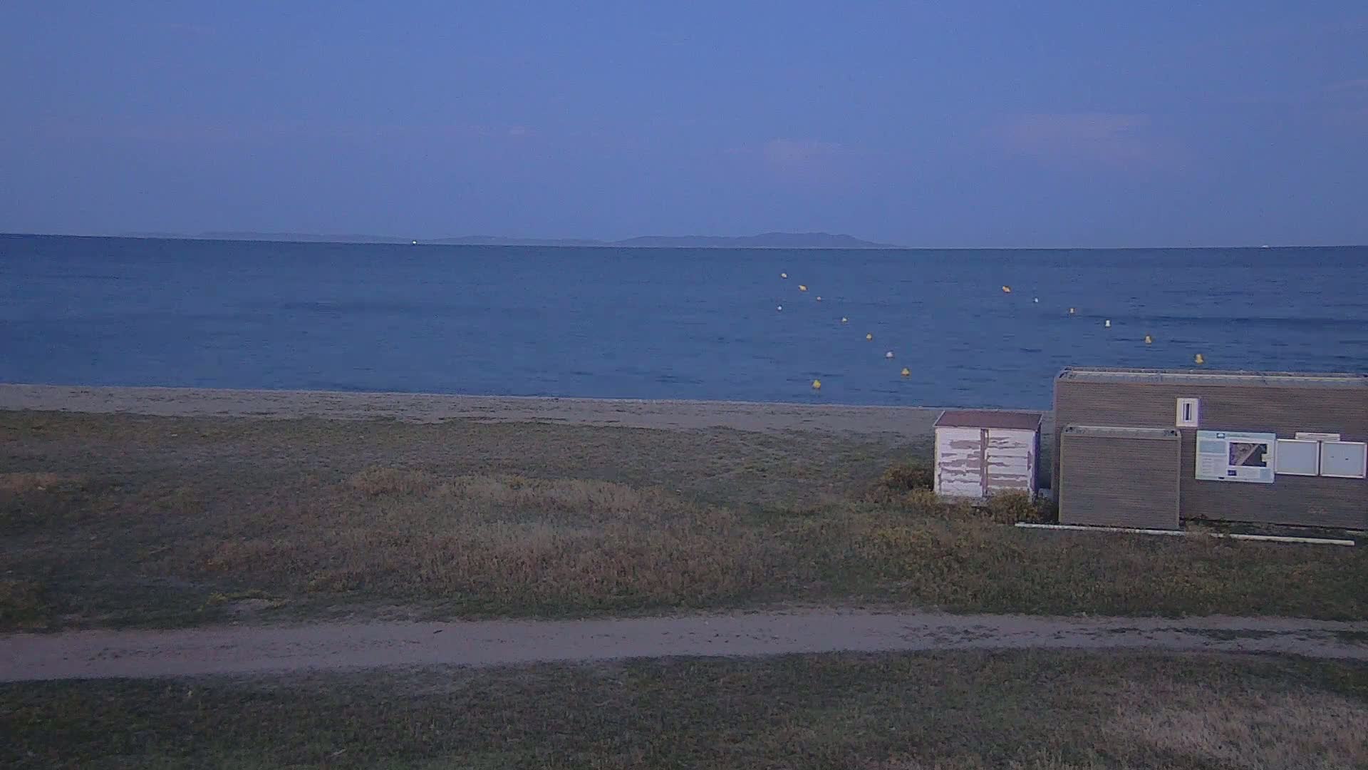A calm, dark-blue sea meets a sandy beach under a twilight sky, with two small buildings situated on the adjacent land.