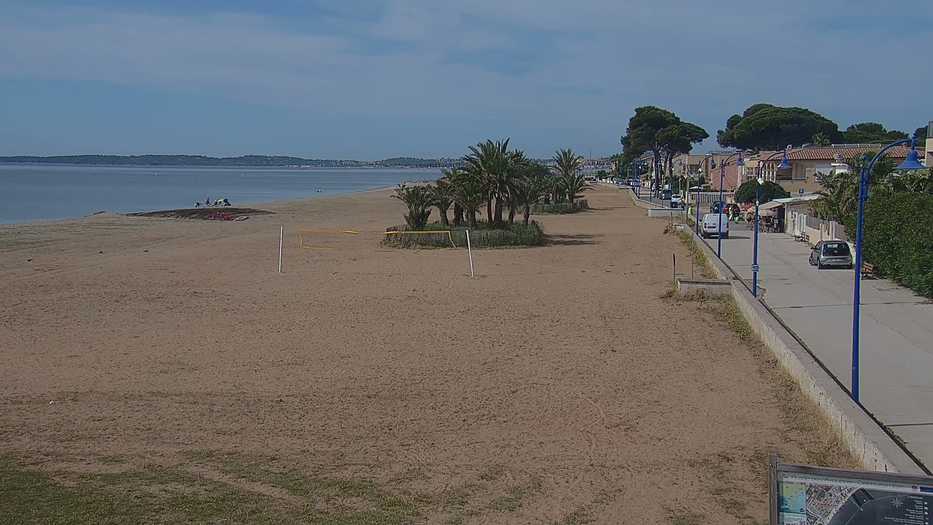 A sandy beach stretches along a calm, blue body of water under a mostly sunny sky, with a paved walkway and buildings lined with palm trees bordering the beach.