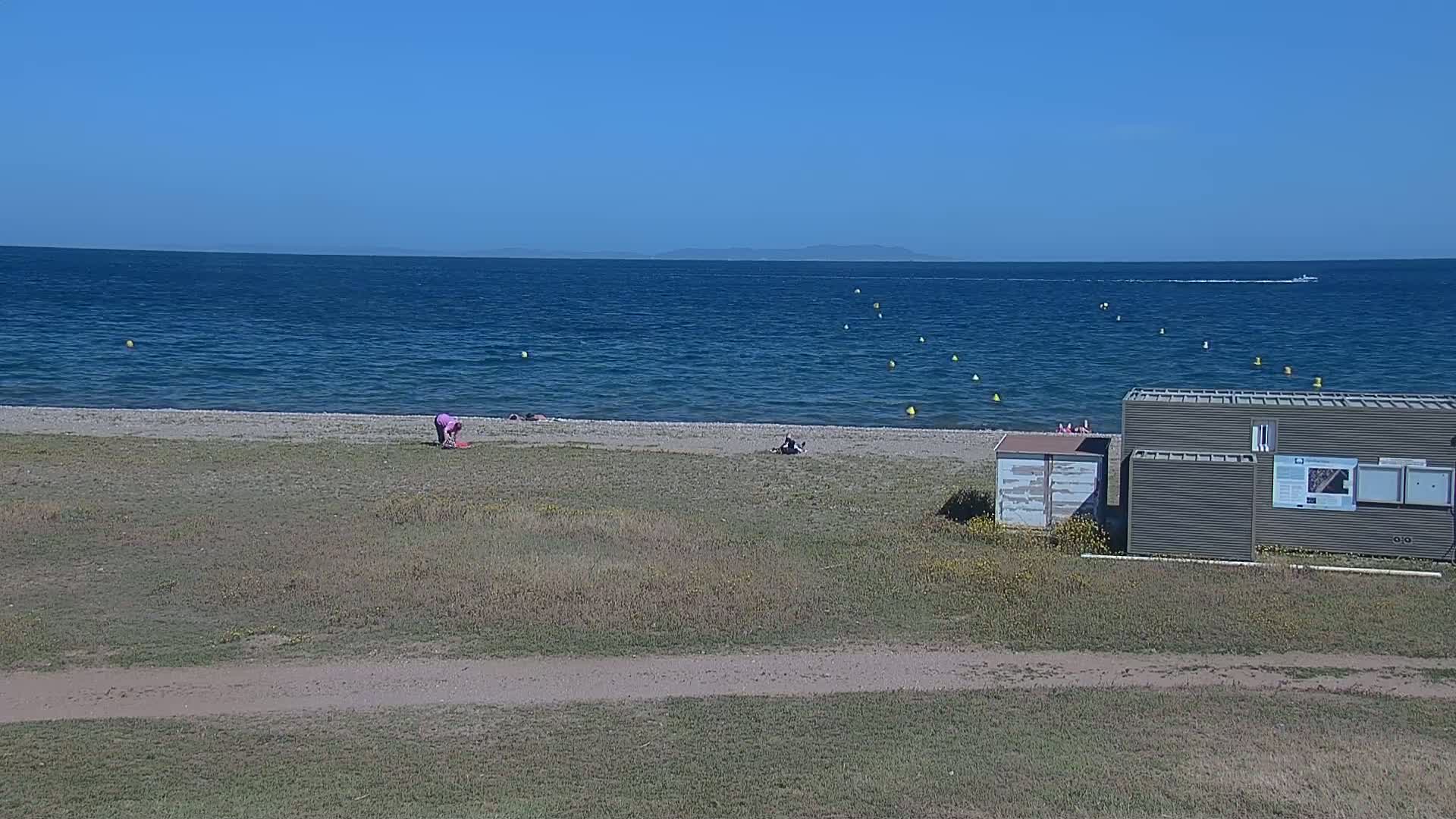 A mostly sunny day at a beach shows a calm sea with a few people on the shore near a small building, with a distant island visible on the horizon.