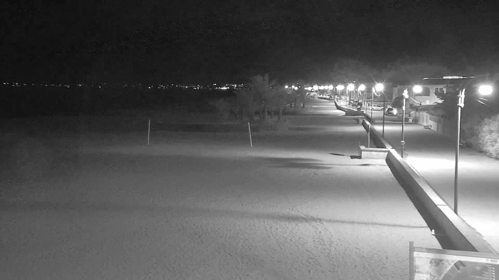A long, sandy beach at night is illuminated by streetlights lining a paved walkway beside it.