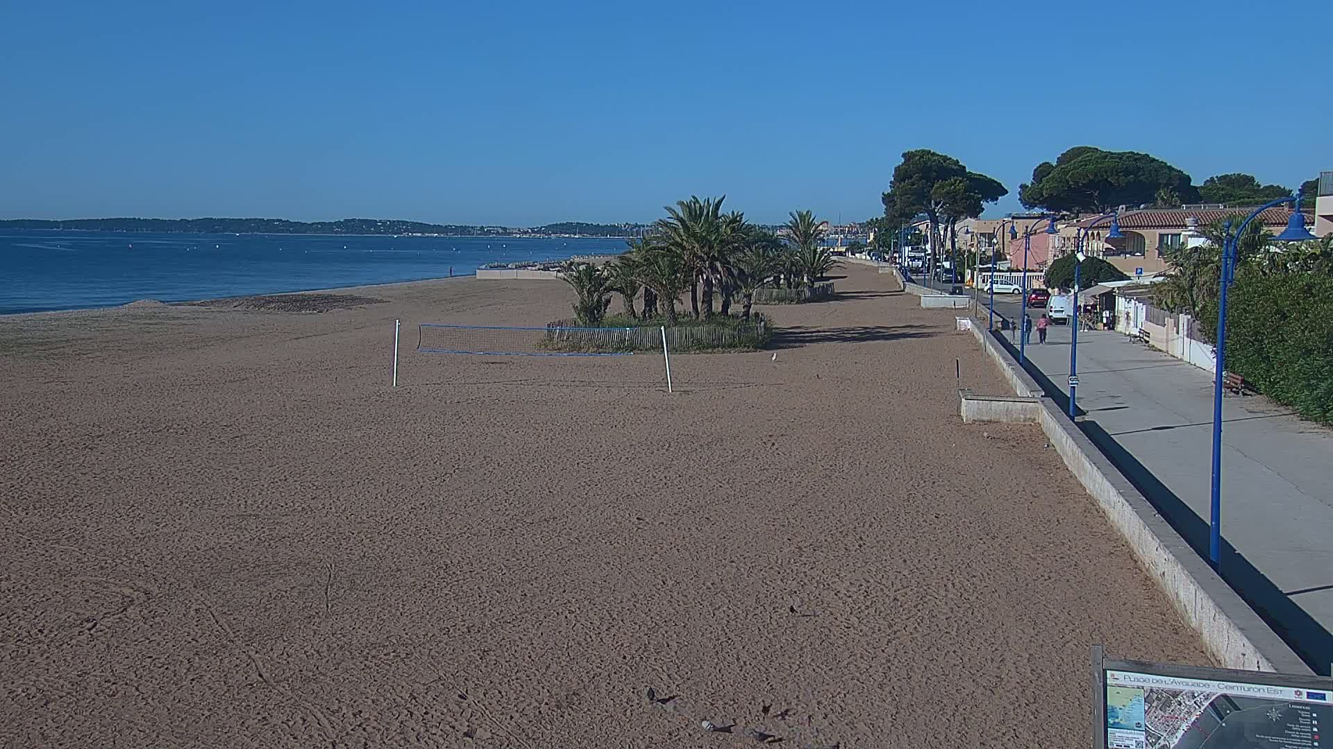 A sandy beach with a volleyball net and palm trees, next to a paved walkway and buildings, on a clear, sunny day.