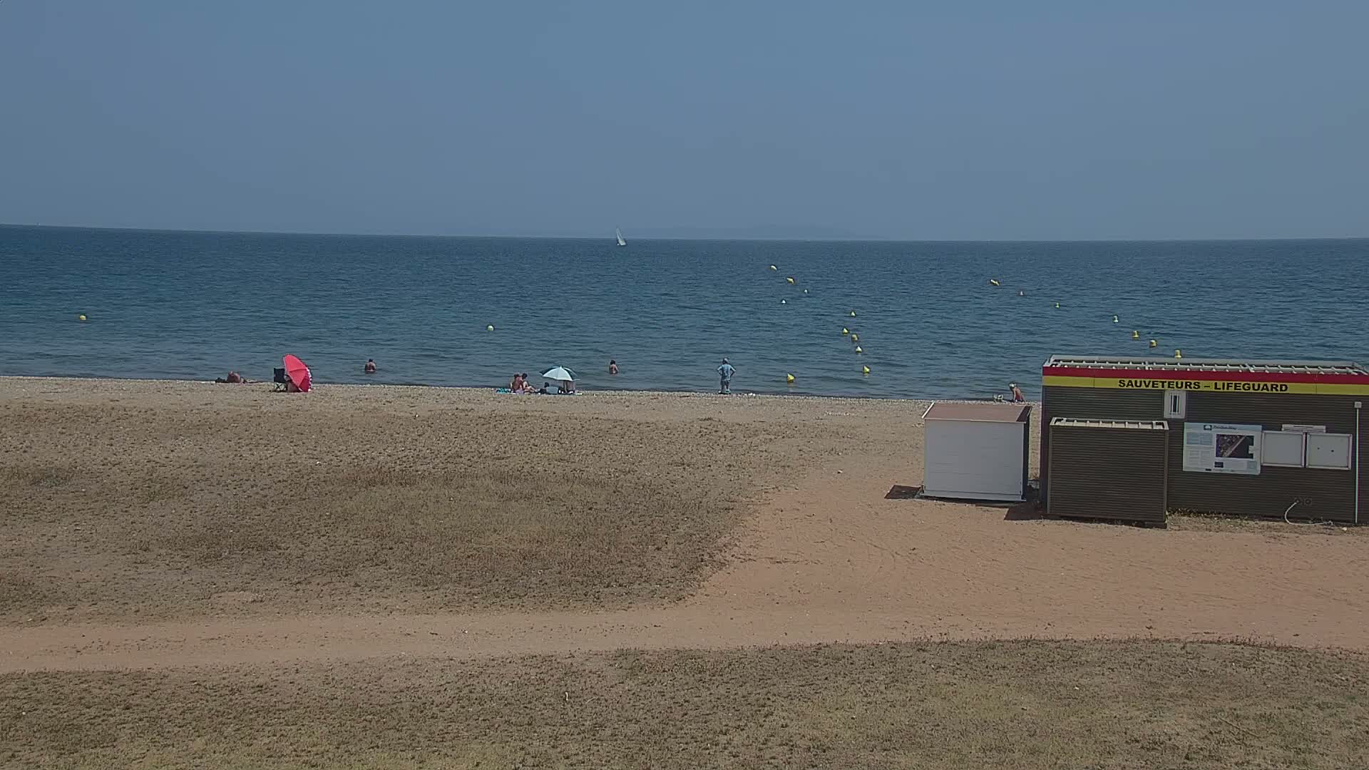 A sandy beach with a few people and umbrellas, a lifeguard station, and a calm blue sea under a clear sky.
