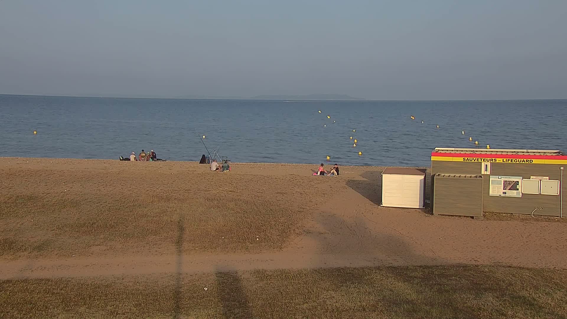A sandy beach with a few people sitting near the water's edge, a lifeguard station, and calm water under a clear sky.