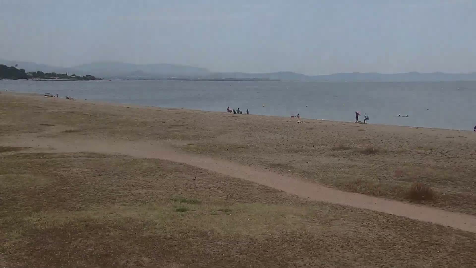 A sandy beach with a few people on it and a calm, grey sea under an overcast sky stretches to a distant shoreline.
