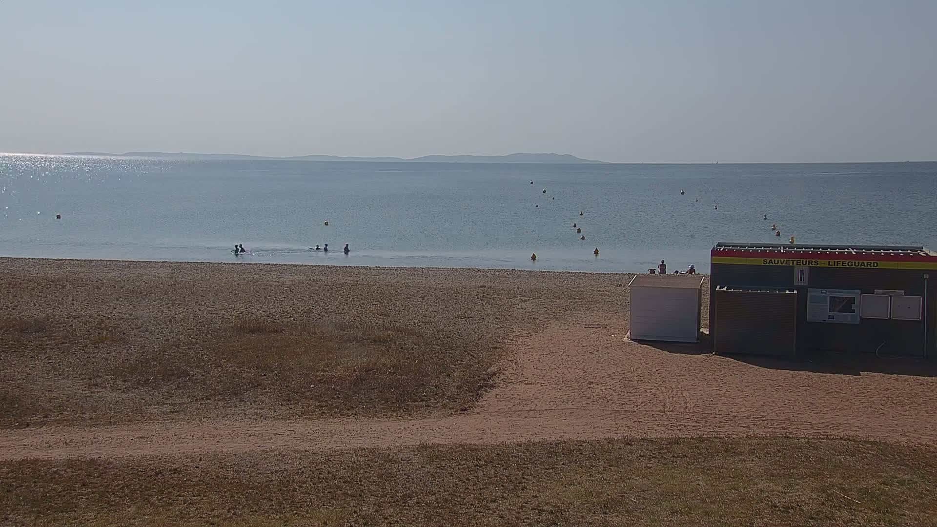 A sandy beach with a calm, light blue sea, a few people in the water, and a lifeguard station under a clear, sunny sky.