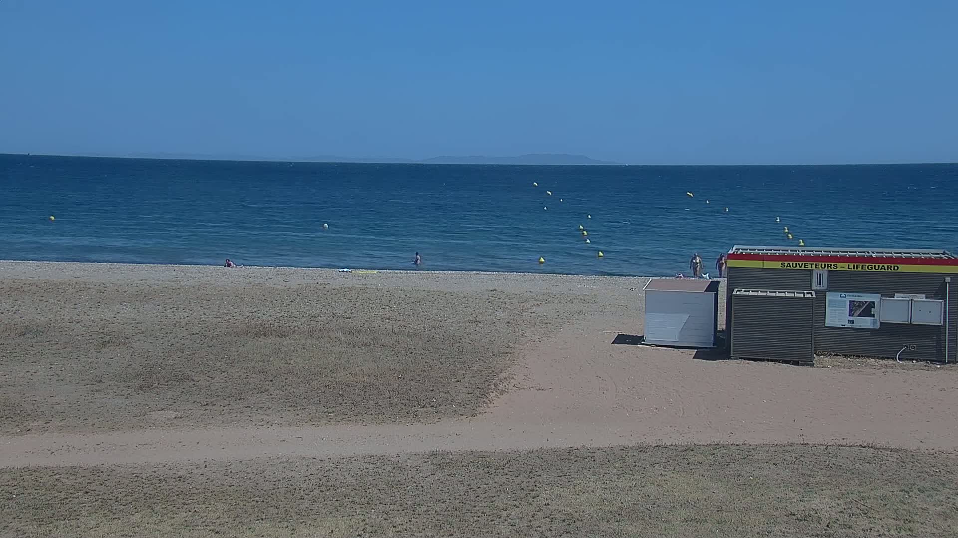 A sandy beach with a few people on it and a lifeguard station is next to a calm blue ocean under a clear sky.