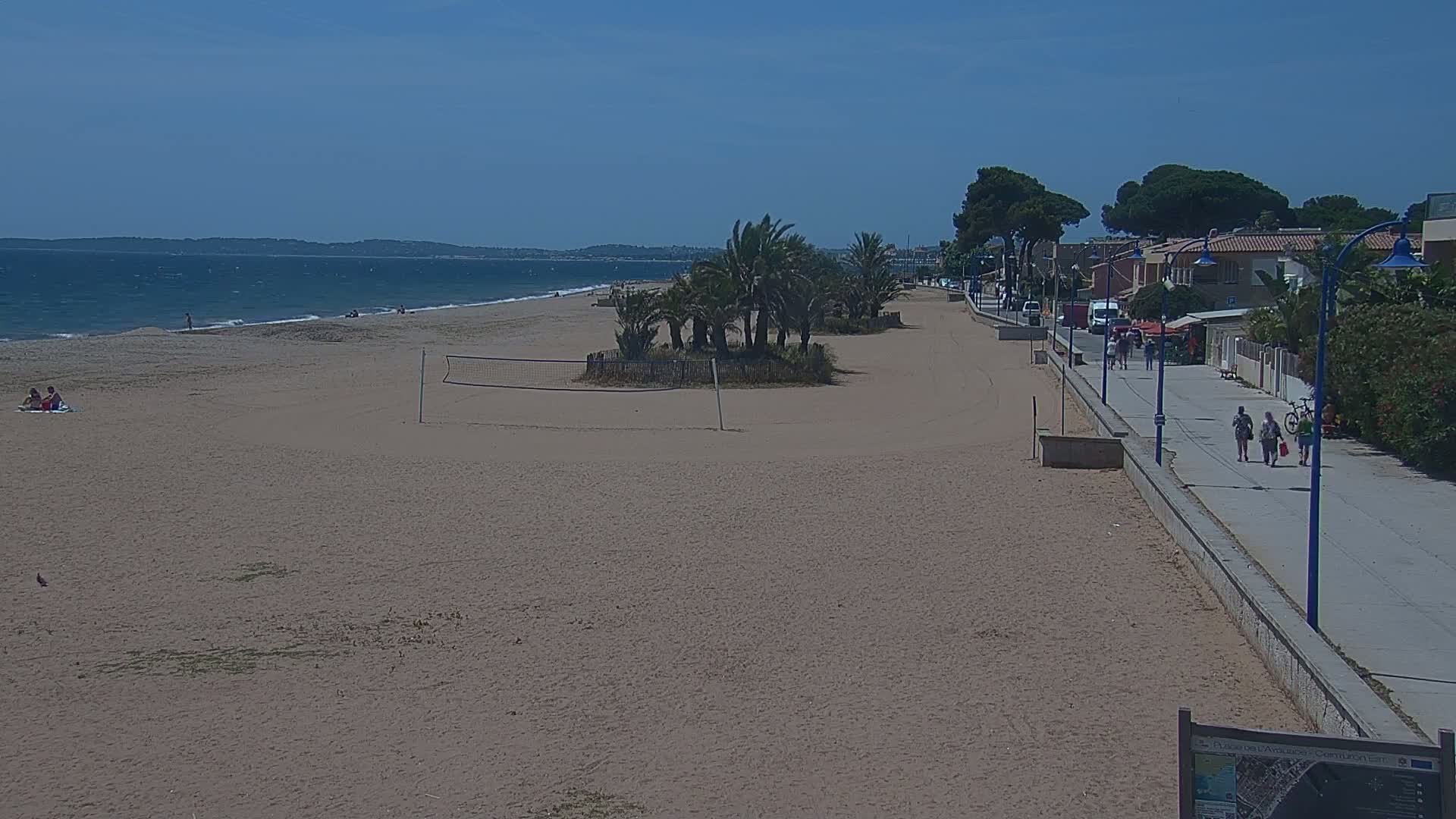 A sandy beach with a volleyball net, palm trees, and a paved walkway leading to buildings is visible under a sunny, clear sky.