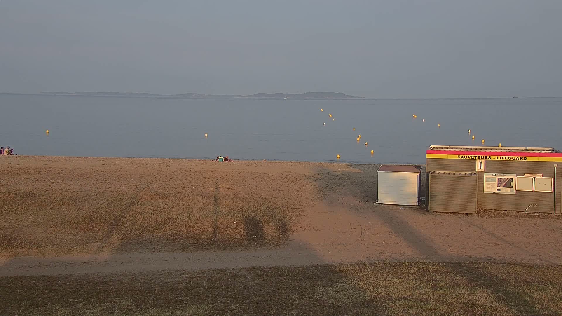 A sandy beach with a lifeguard station, a few people, and yellow buoys in a calm, gray-blue sea under a clear, pale sky.