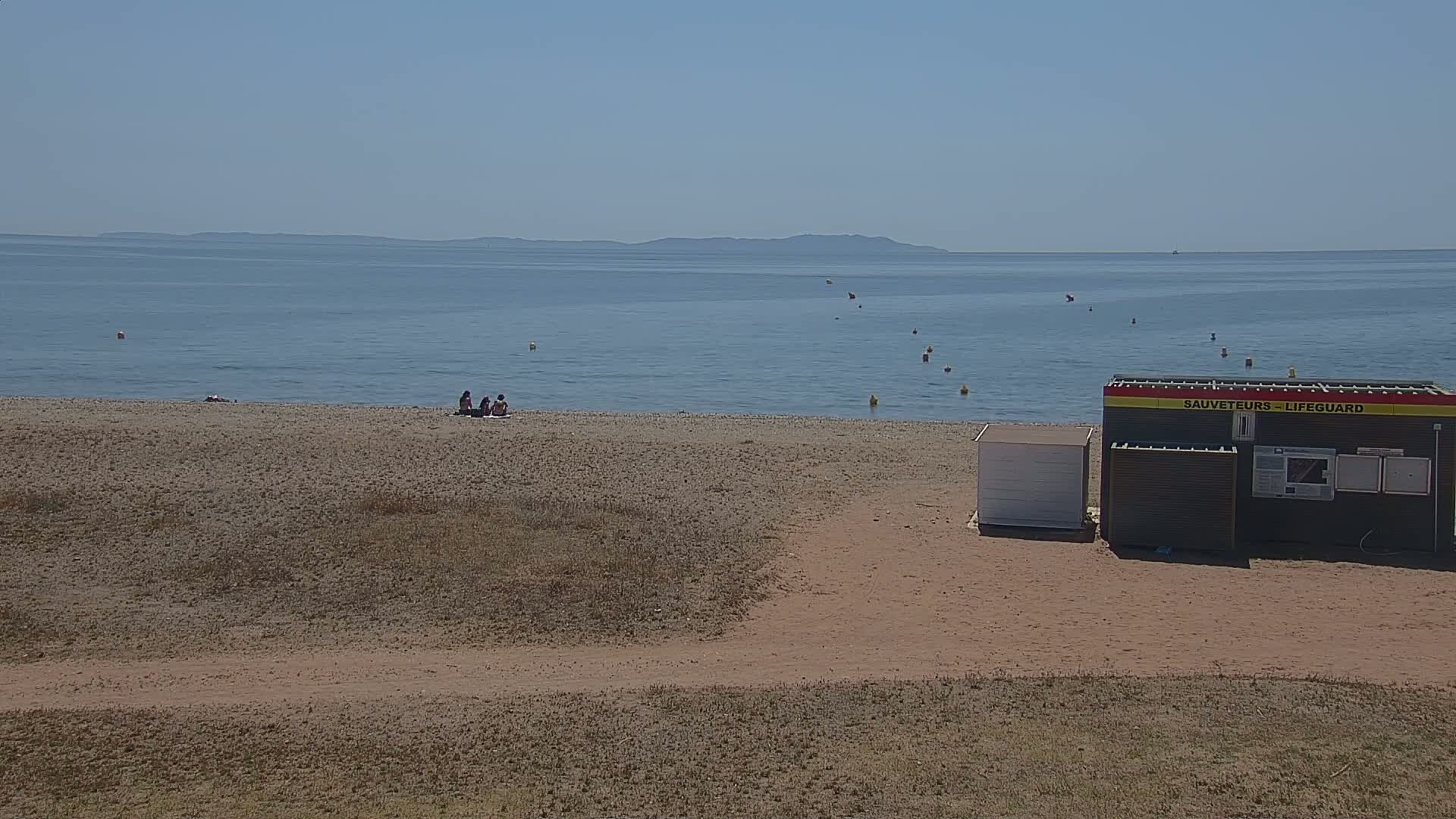 A sandy beach with a few people sitting near calm blue water under a clear sky, and a lifeguard station nearby.