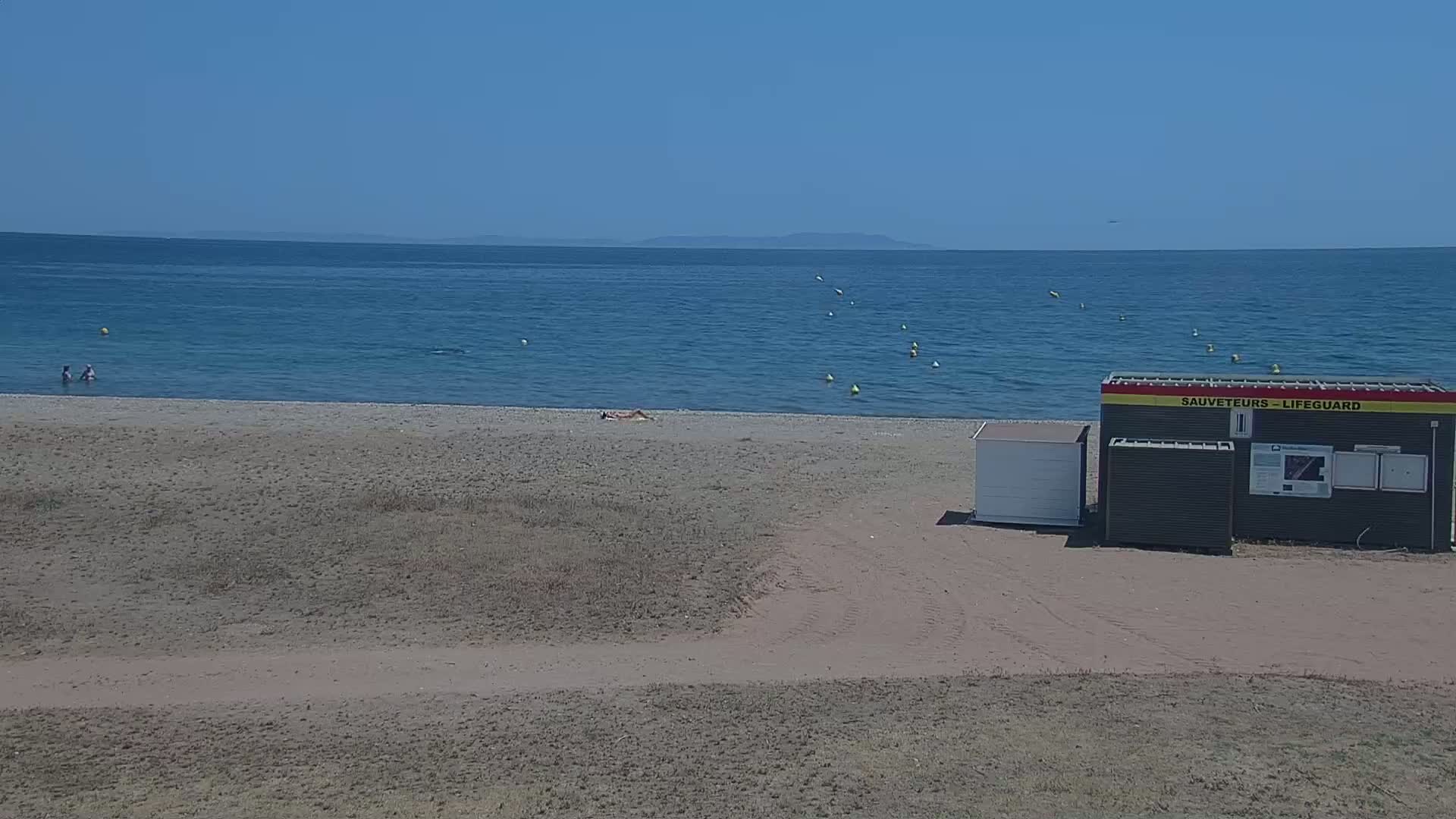 A sandy beach with a few people, a lifeguard station, and a calm blue ocean under a clear sky.