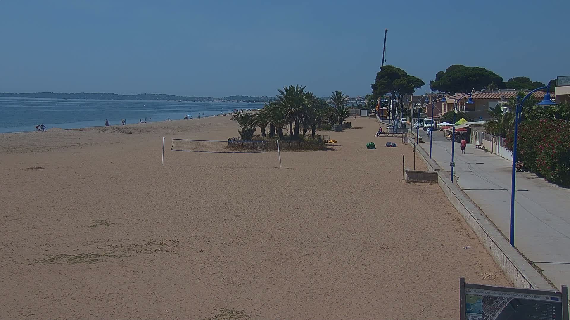 A sandy beach with a volleyball net and palm trees stretches to a calm sea under a clear blue sky.
