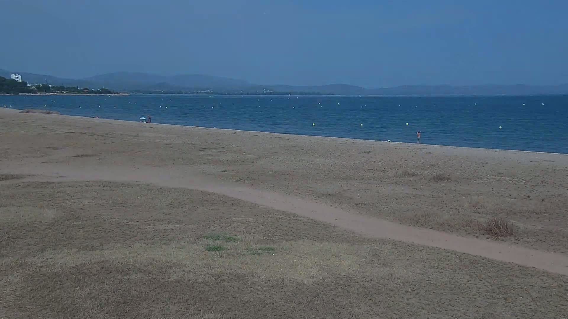 A mostly empty sandy beach stretches toward a calm blue sea under a clear sky, with a few people visible on the sand and in the water, and distant mountains in the background.
