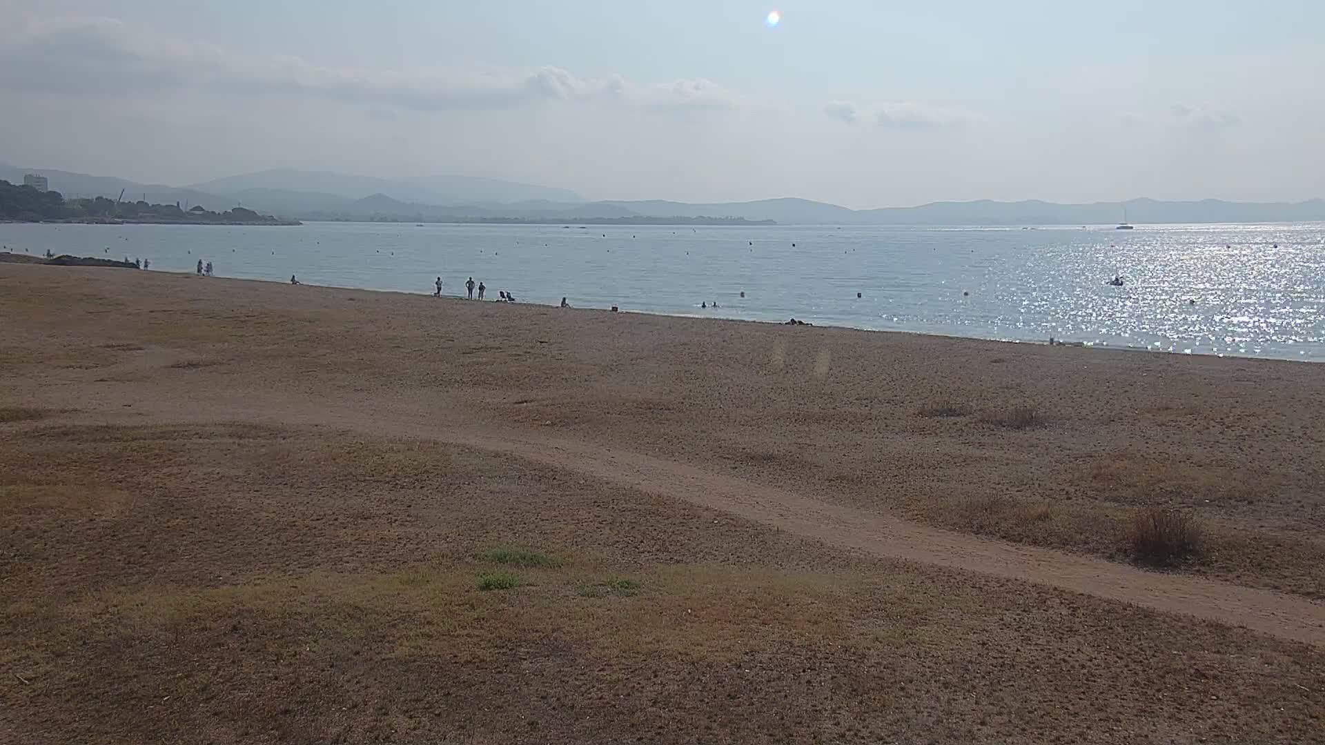 A sandy beach with a few people on it stretches along a calm, sunlit sea under a mostly clear sky, backed by distant mountains.