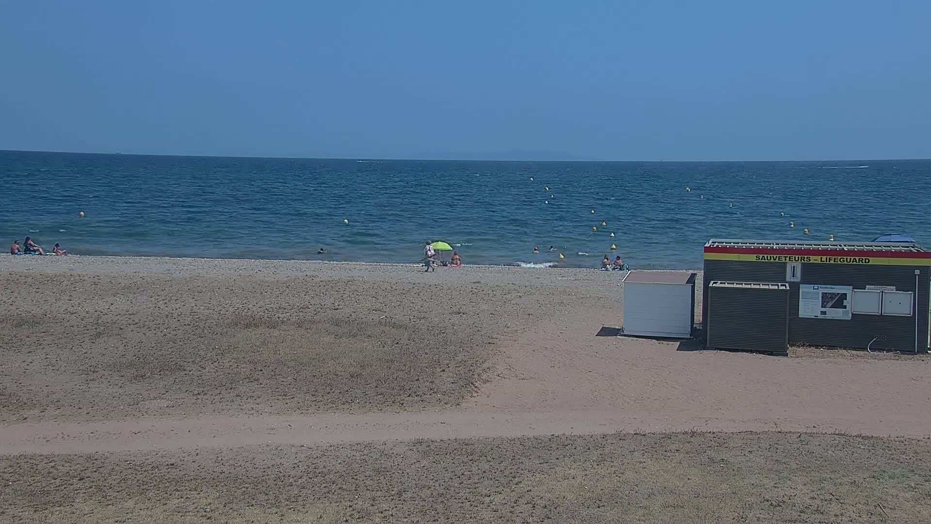 A sandy beach with a few people sunbathing and swimming in a calm, blue sea under a clear sky, next to a lifeguard station.
