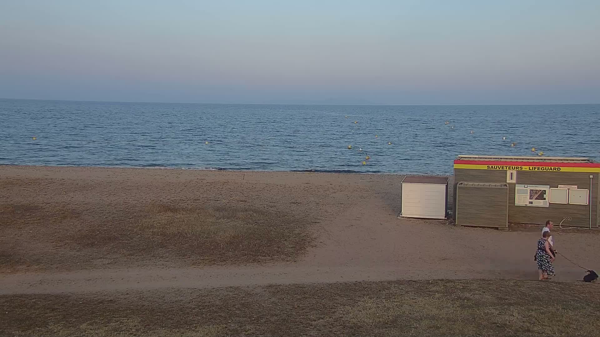 A calm, twilight ocean meets a sandy beach where two people walk a dog past a lifeguard station.