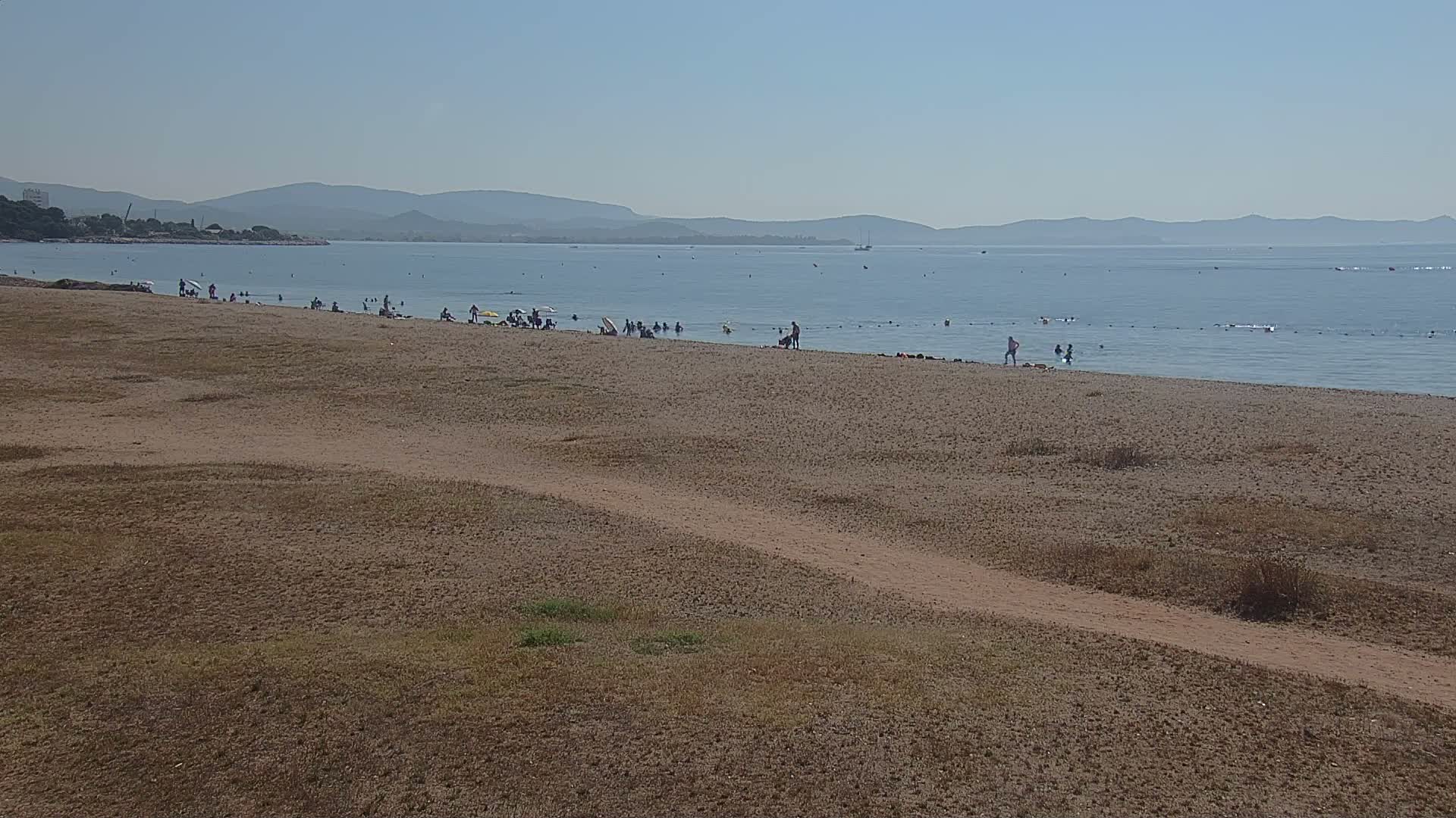 A sandy beach with scattered people enjoying the calm, clear water under a bright, sunny sky, backed by distant mountains.