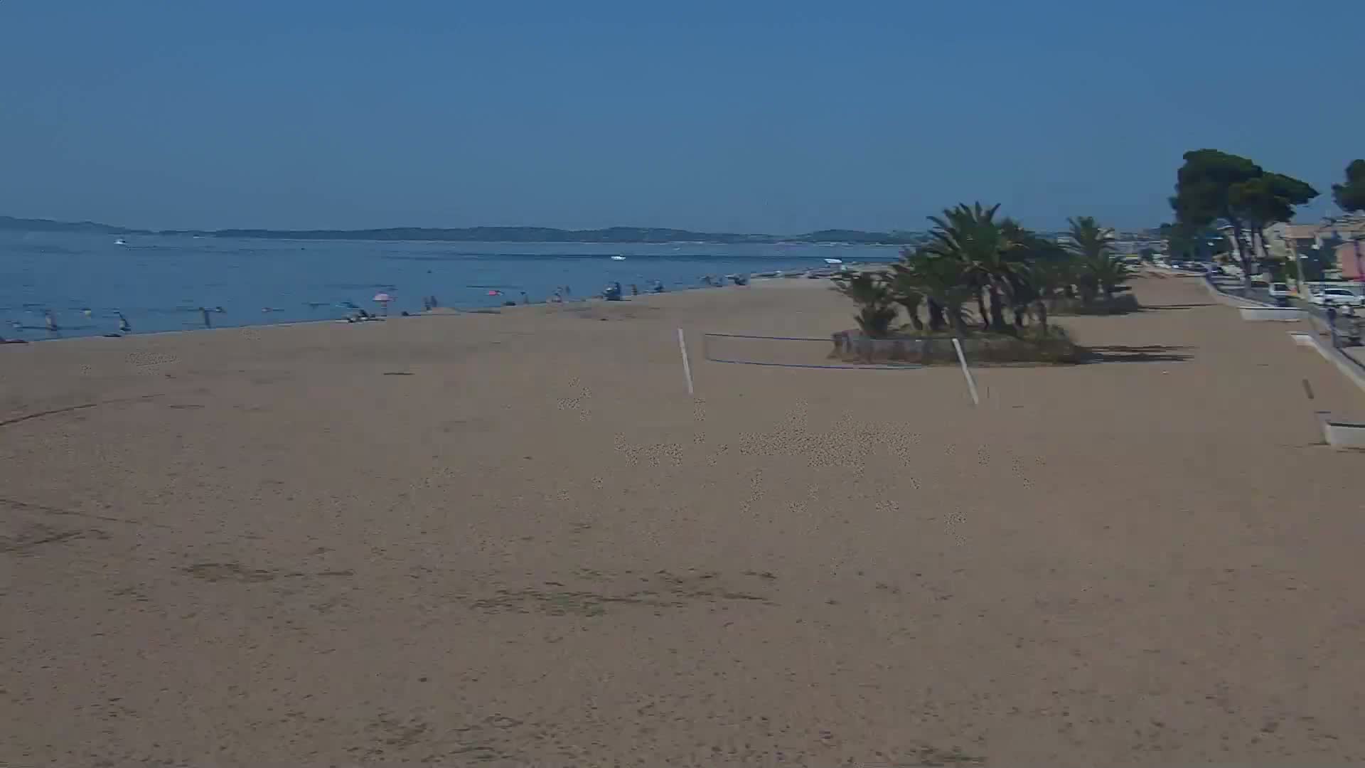 A sandy beach with a few people on it stretches towards a calm sea under a clear blue sky, lined with palm trees and a paved walkway.