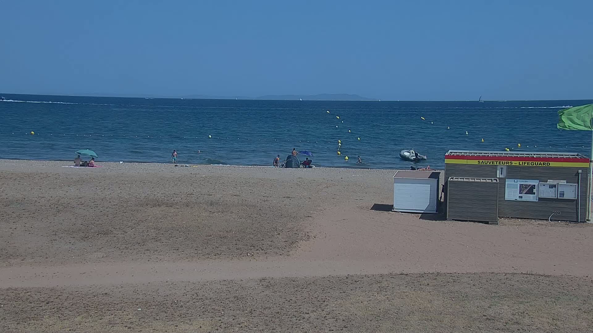 A sandy beach with a few people and a lifeguard station is visible on a sunny day, with calm blue water extending to a distant coastline.