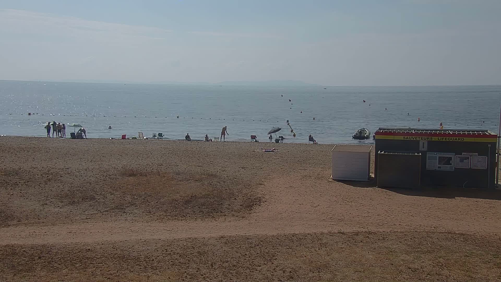 A sandy beach with a few people swimming and relaxing near a lifeguard station on a calm, sunny day.
