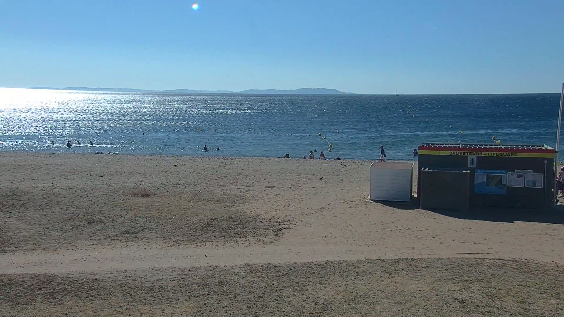 A sandy beach with a few people swimming and relaxing near a lifeguard station, under a clear blue sky.