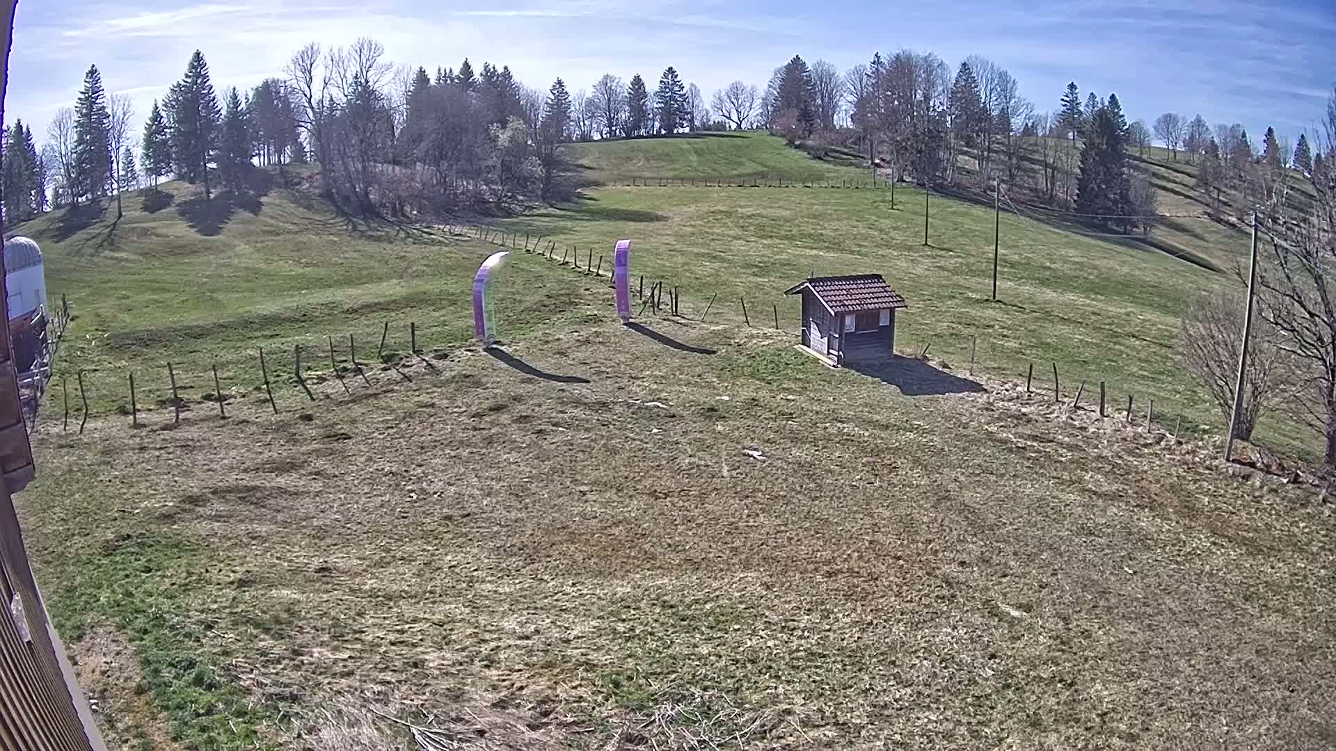 A grassy hillside under a sunny sky features a small wooden shed, two tall purple banners, and a wooden fence.
