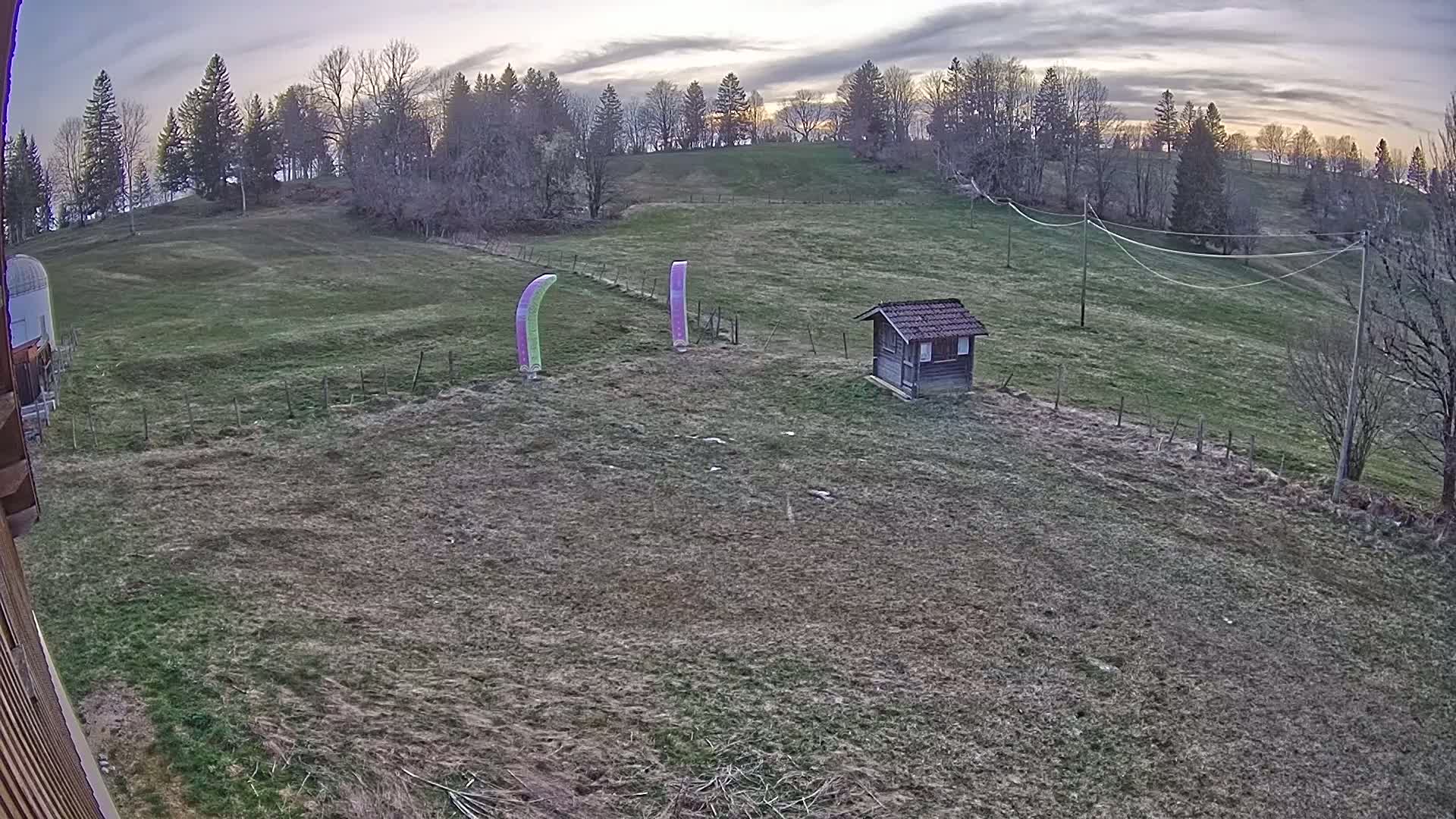 A grassy field under a cloudy sunset sky contains two tall, slender, vertical banners and a small wooden shed near a treeline.