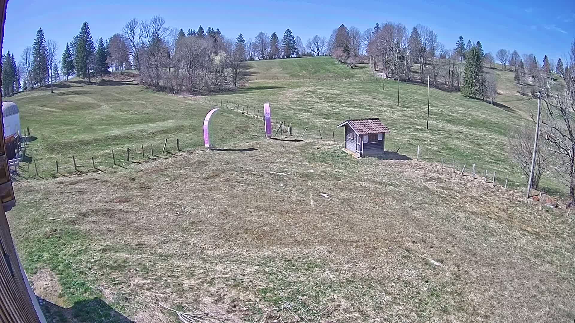 A grassy hillside under a clear blue sky features two tall pink arches and a small wooden shed near a short fence.
