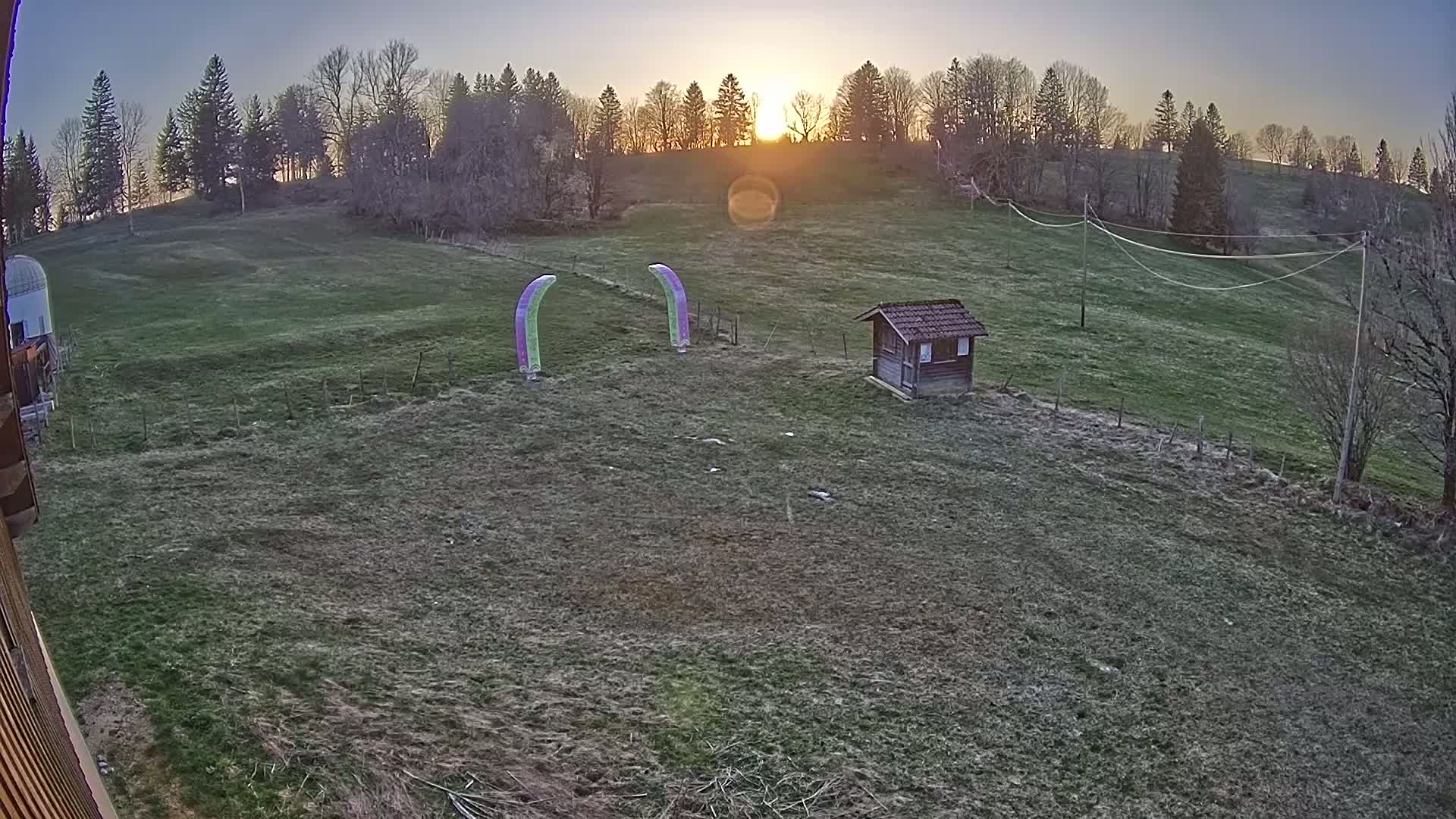 A grassy field at sunset, with a small wooden shed and two arched markers, is surrounded by a line of trees.