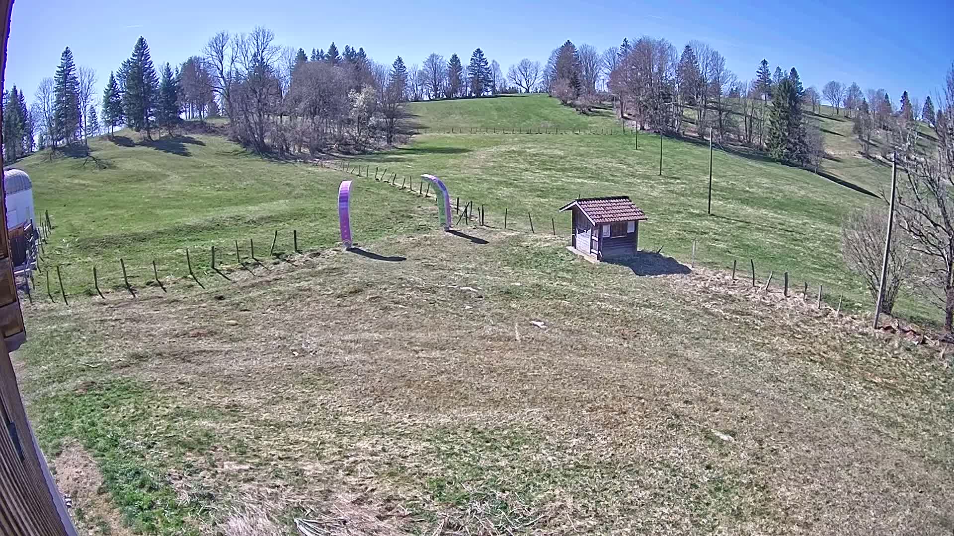 A grassy field under a clear blue sky contains a small wooden structure, two curved purple and green structures, and a short wooden fence.