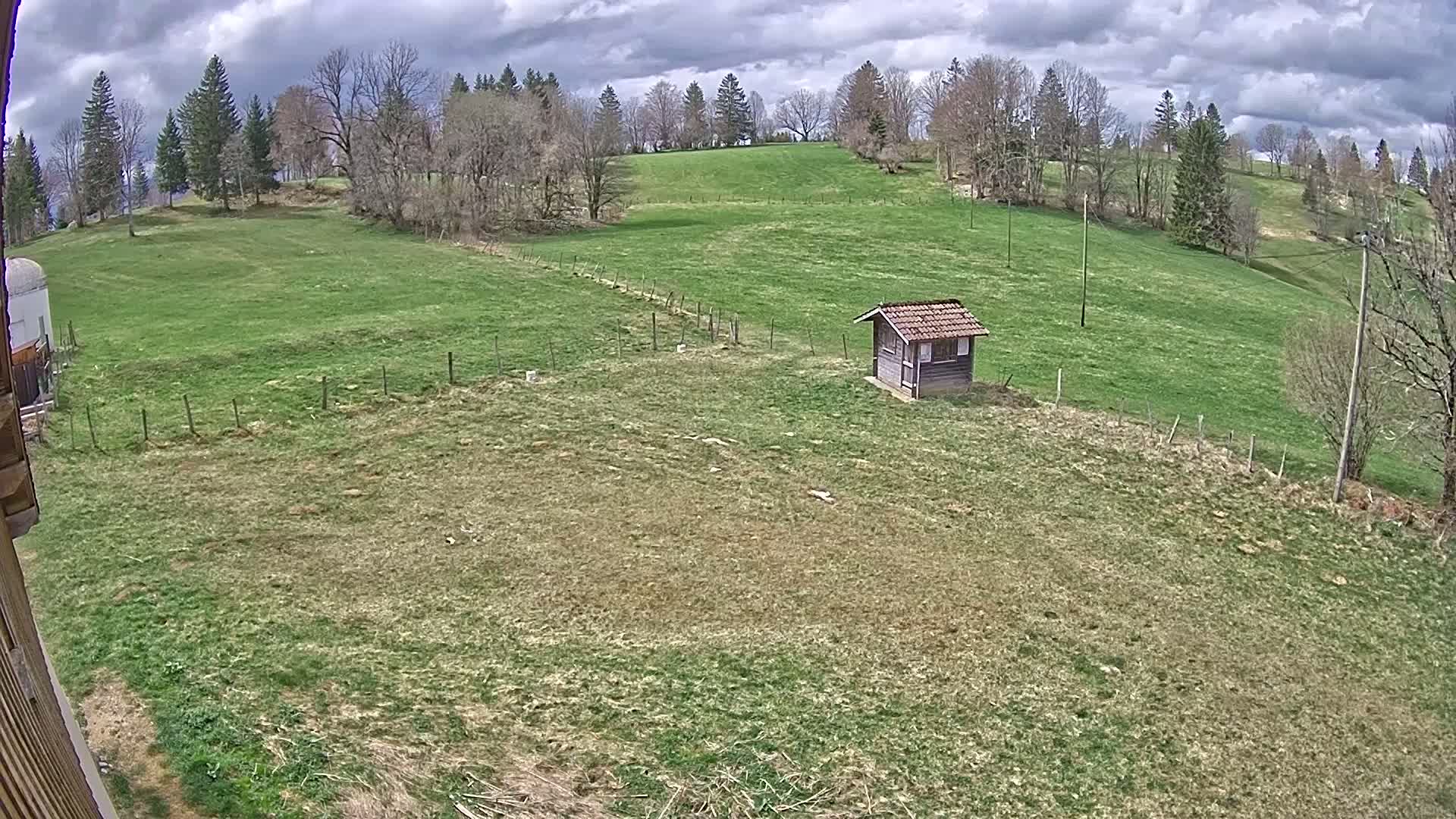 A small wooden shed sits in a grassy field under a cloudy sky, surrounded by trees and a wire fence.
