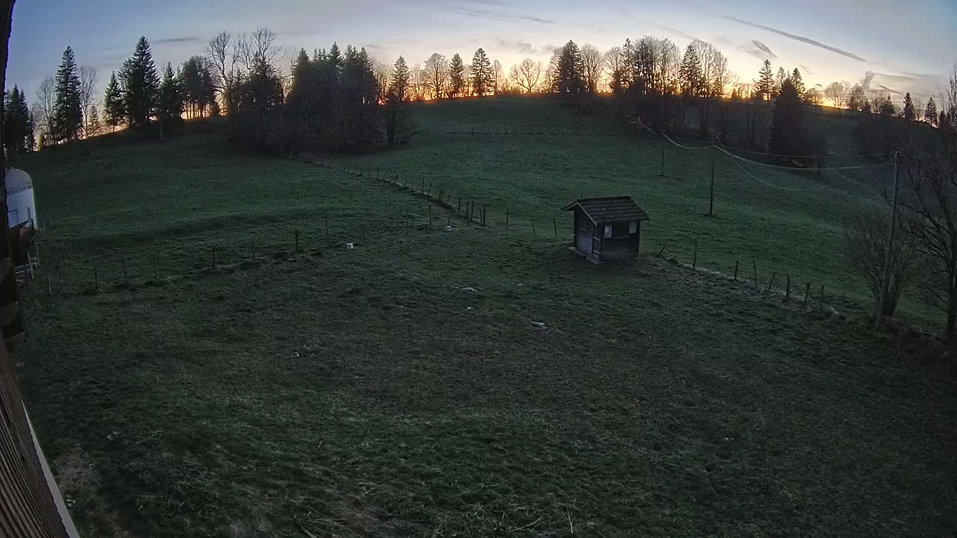 A small shed sits in a grassy field at dusk, with a line of trees silhouetted against a twilight sky.