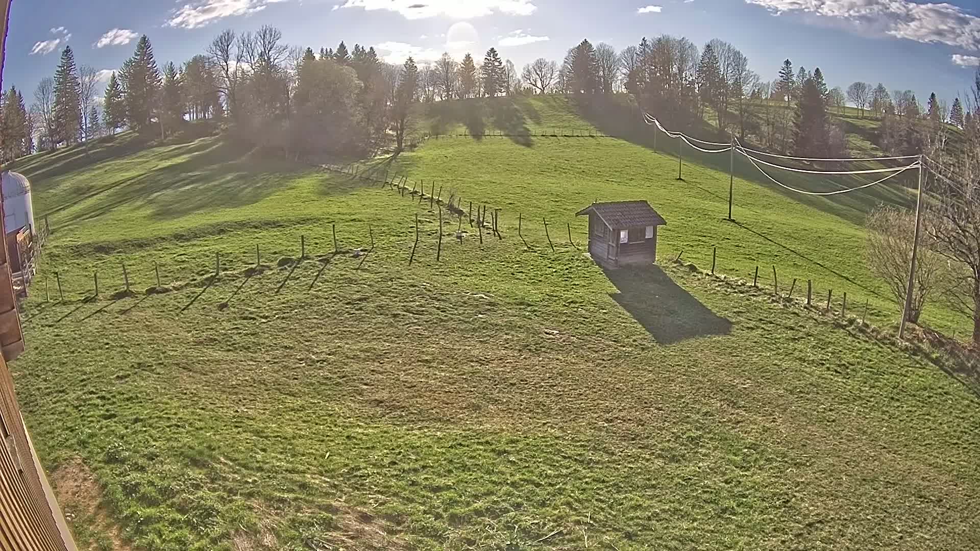 A small wooden shed sits in a grassy field bordered by a low fence, under a mostly sunny sky with some clouds.