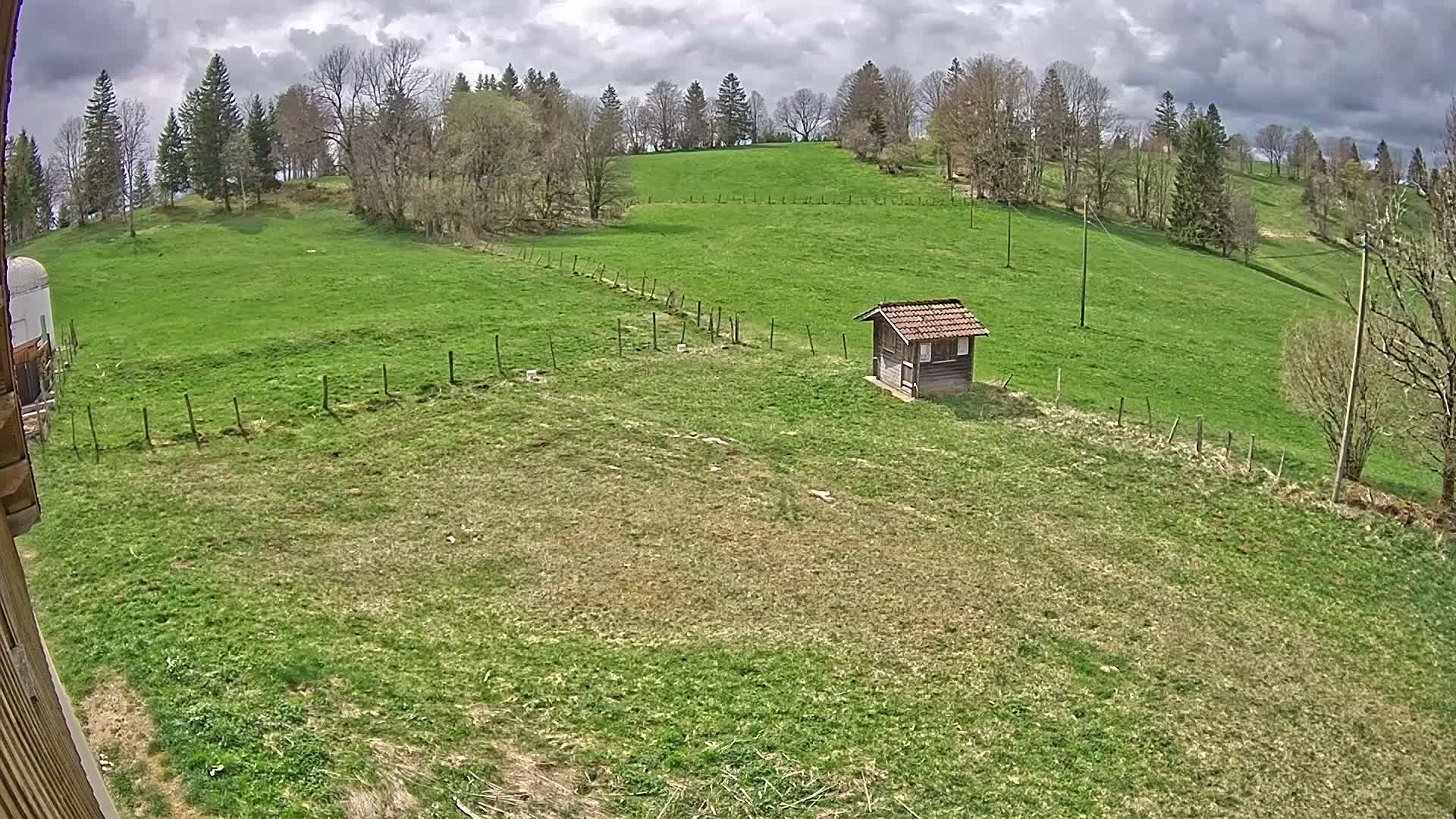 A small wooden shed sits in a sprawling green field under a partly cloudy sky, bordered by a wooden fence and surrounded by trees on a hillside.