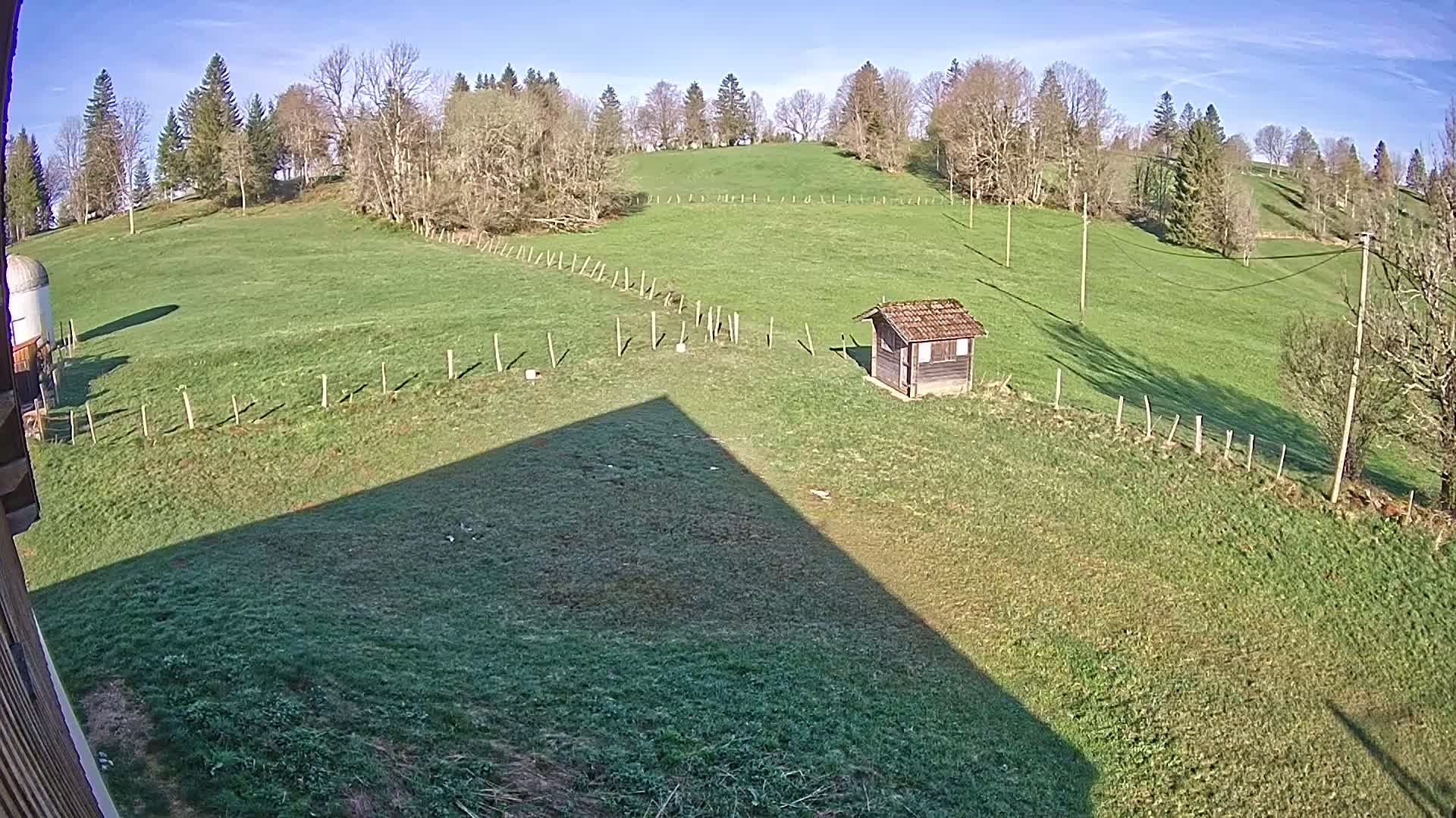 A grassy hillside with a small shed, under a clear blue sky, is partially shaded by a long, sharp shadow.