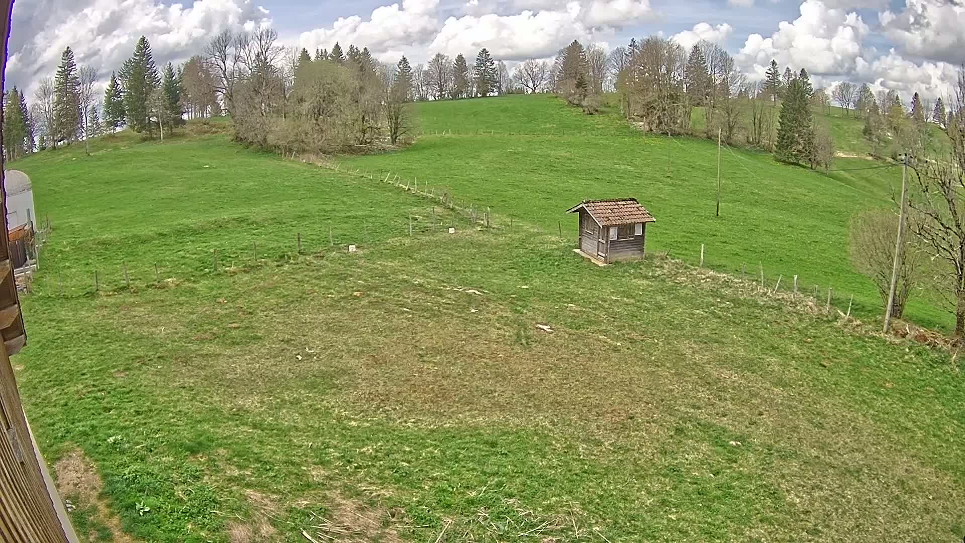 A small wooden shed sits in a grassy field under a partly cloudy sky, surrounded by trees on a hillside.