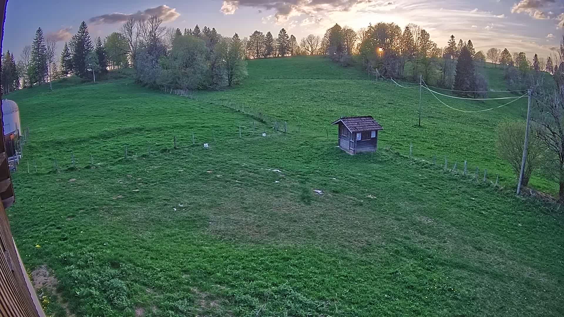 A small wooden shed sits in a large green field, surrounded by trees under a partly cloudy sunset sky.